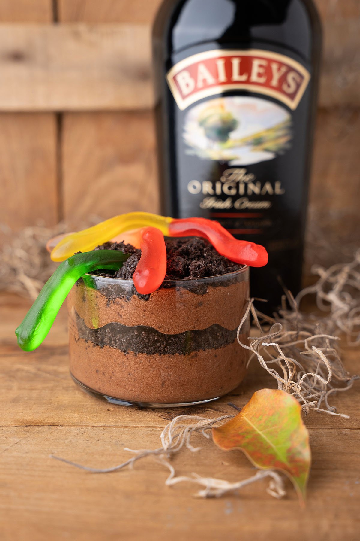 A Boozy Dirt Cup dessert with gummy worms on top sits in front of a Baileys bottle on a wooden surface.