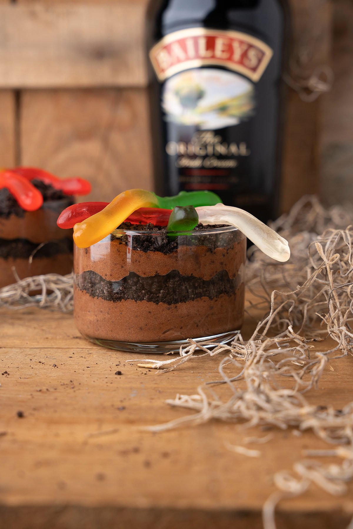 A glass of Boozy Dirt Cups chocolate mousse with gummy worms on top, set on a wooden table with a Baileys bottle in the background.