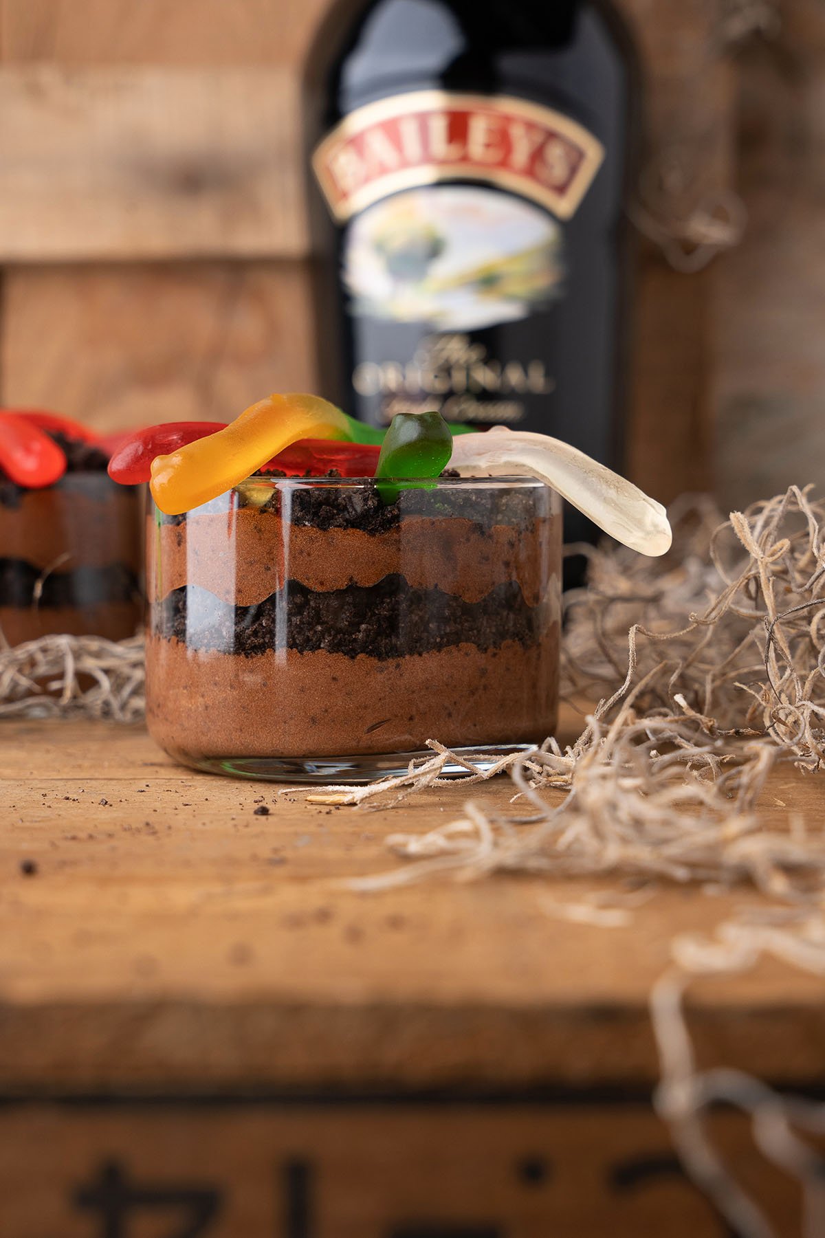 Chocolate mousse in a glass, topped with gummy worms for a playful twist on dirt cups for adults, with a Baileys bottle blurred in the background.
