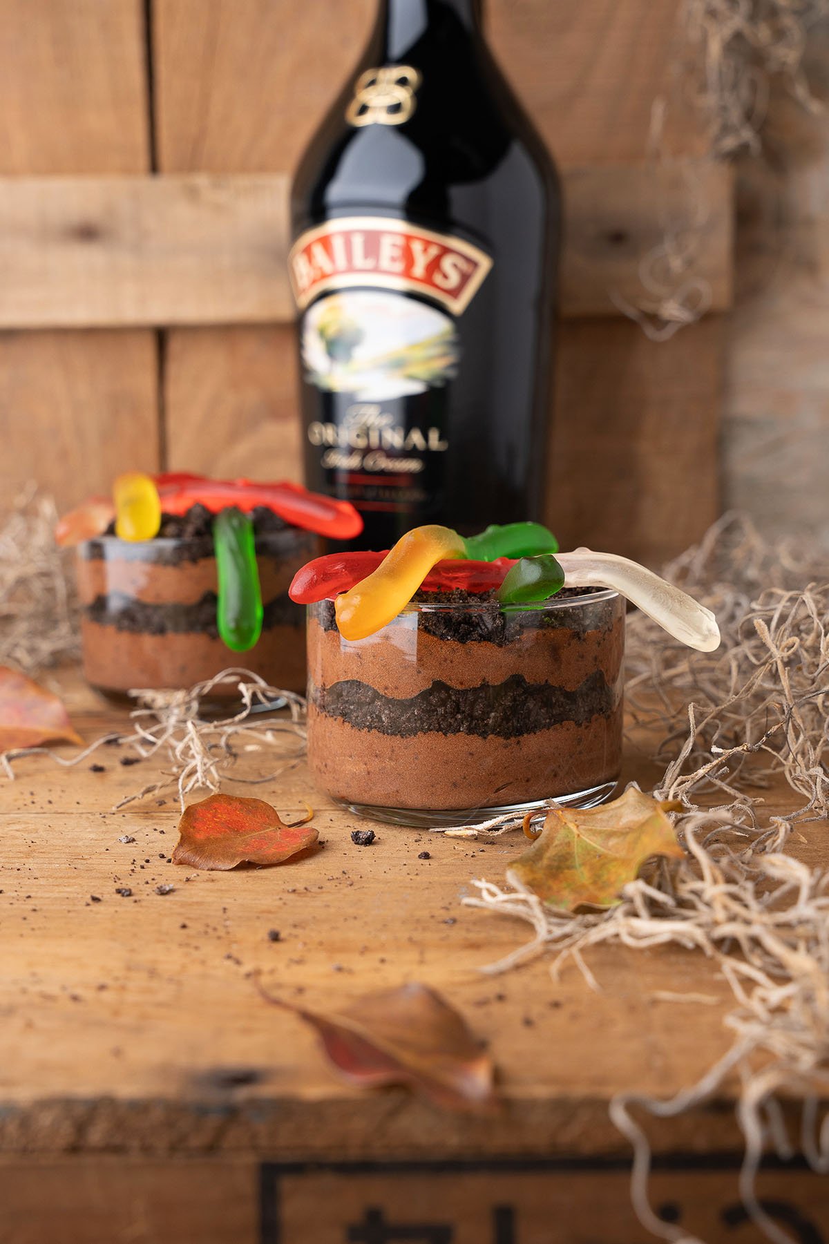 Chocolate mousse cups topped with gummy worms, with a bottle of Baileys and autumn leaves in the background—these Boozy Dirt Cups are a fun, grown-up twist on classic worm and dirt parfaits.