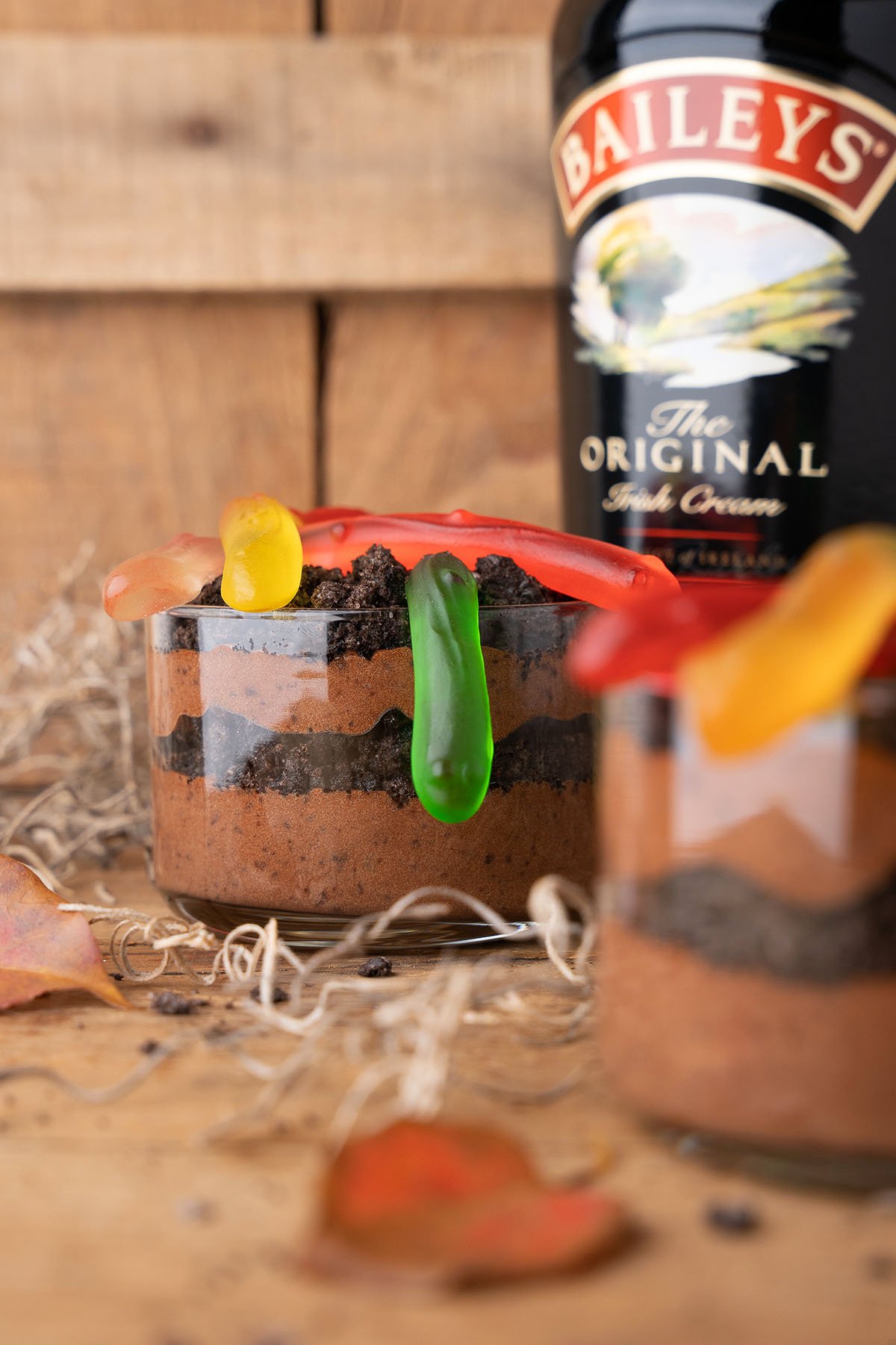 Chocolate mousse in a glass, topped with crushed cookies and gummy worms, creates irresistible dirt cups for adults—Baileys bottle in the background hints at these boozy dirt cups.