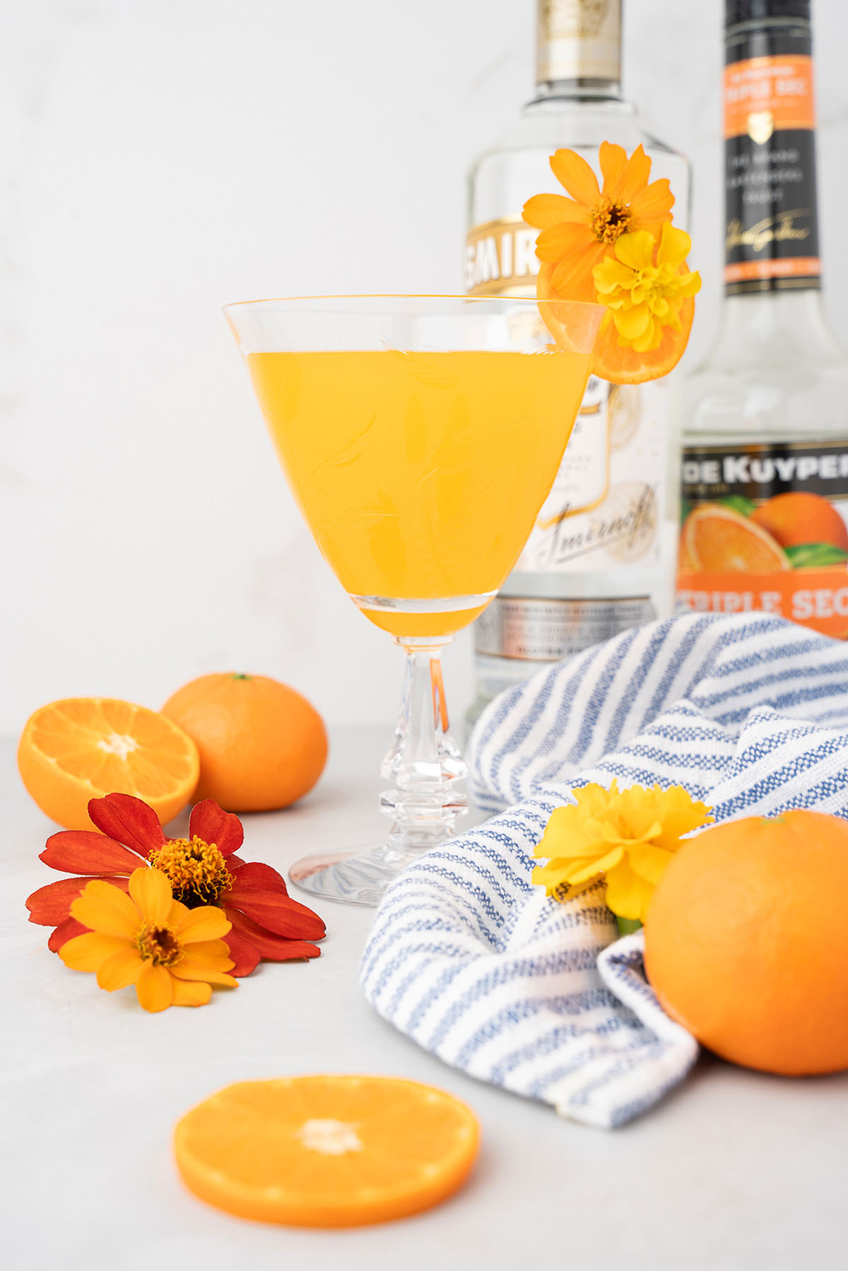 A bright orange summer cocktail, crafted with whipped cream flavoured vodka and triple sec, is garnished with a yellow flower and surrounded by oranges, flowers, and liquor bottles.