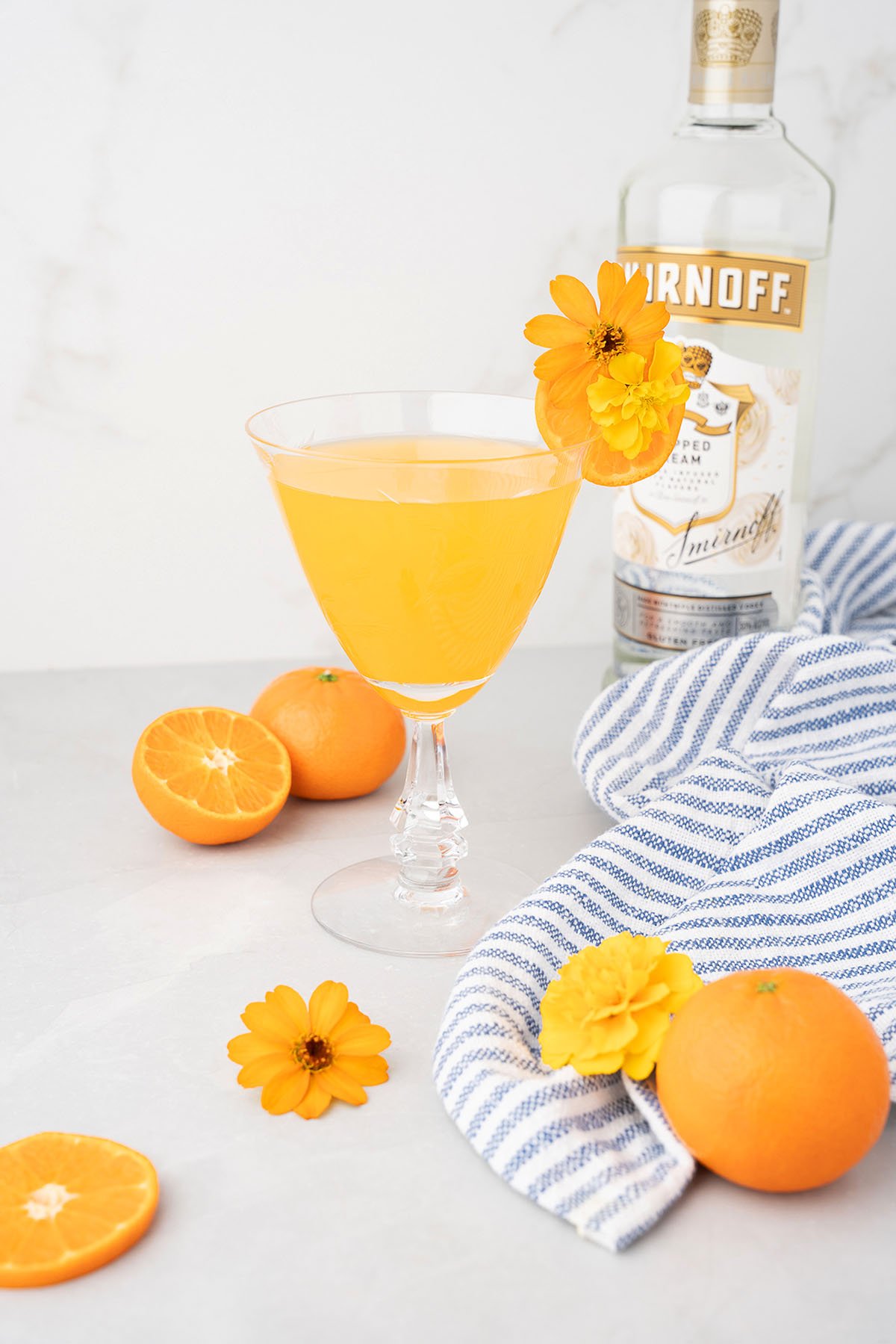 A Clementine Martini garnished with an orange flower, surrounded by fresh oranges and a Smirnoff vodka bottle, is the ultimate Summer Cocktail.