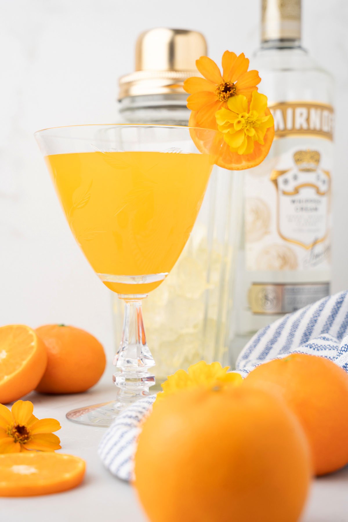 A bright orange summer cocktail, garnished with a marigold flower, sits surrounded by fresh oranges and a vodka bottle—perfect for sipping this season, like an orange dreamsicle martini.