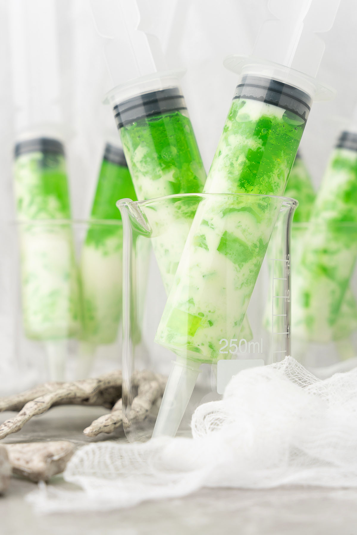 Several syringes filled with green and white layered liquid, perfect for Ectoplasm Syringe Vodka Jello Shots, are placed upright in a clear 250ml beaker.