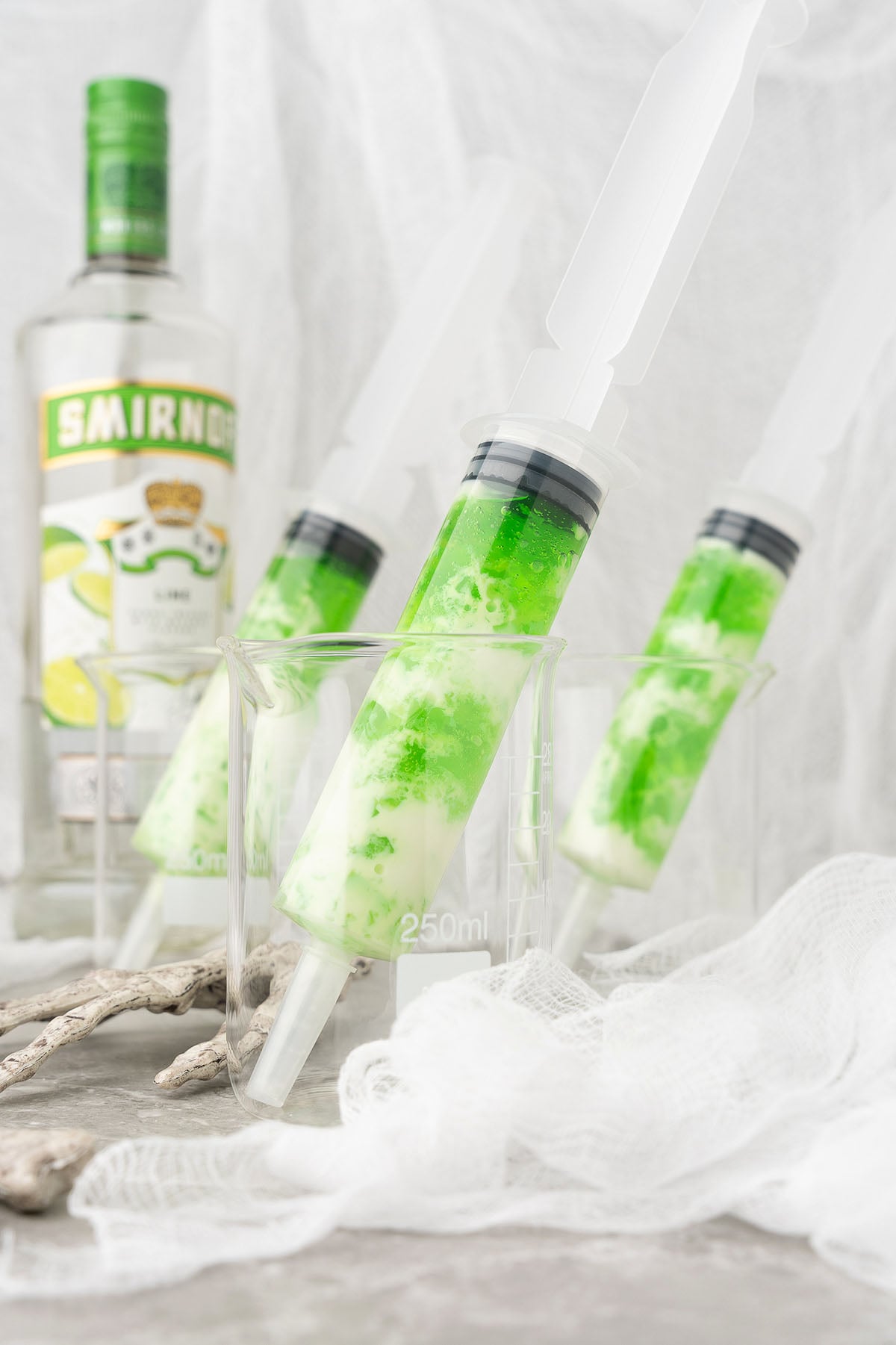 Three green and white cocktail syringes in glass beakers with a bottle of Smirnoff lime vodka in the background—perfect for creative Halloween jello shots or if you want to make jello shots in syringes for your next party.