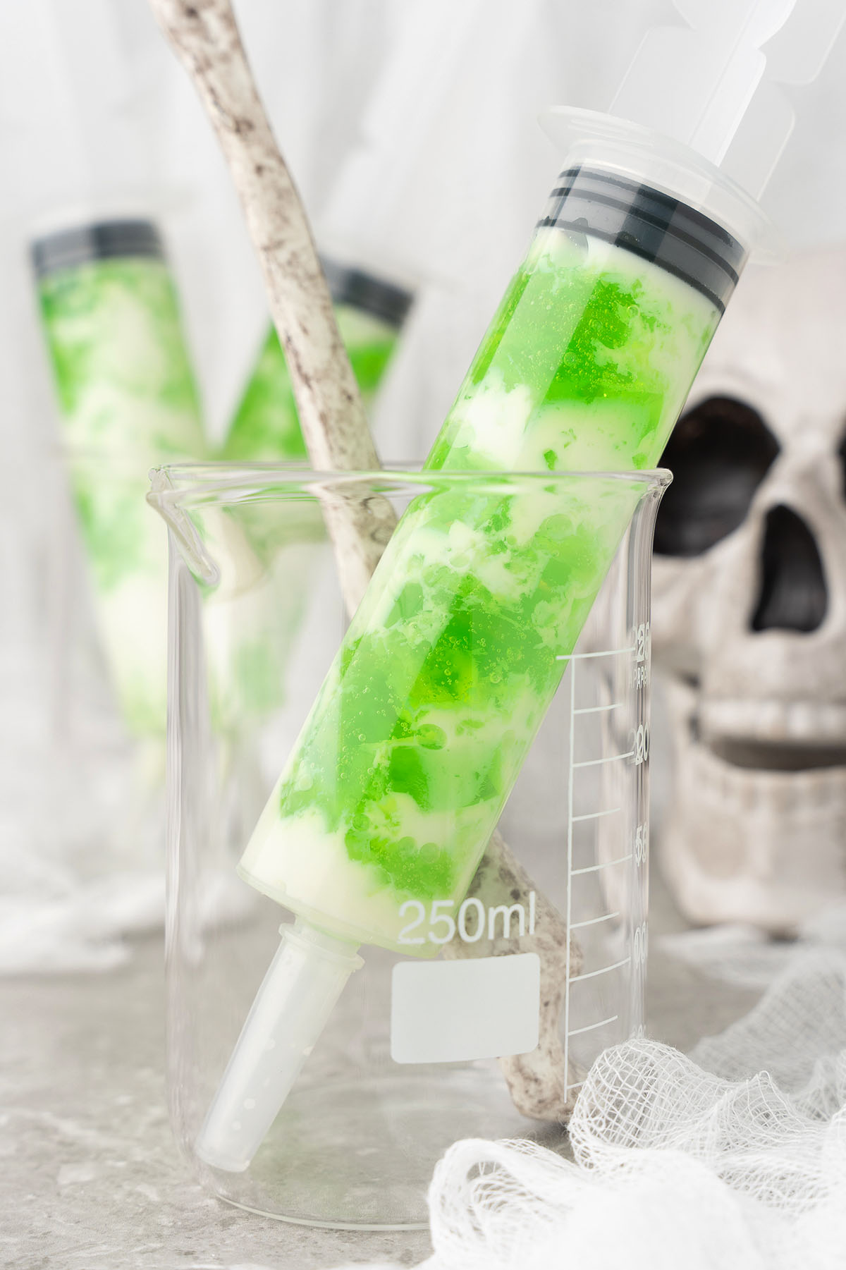 Green and white layered dessert in a syringe, placed in a beaker with a skull decoration in the background—these halloween jello shots are made with Smirnoff lime vodka for a fun and spooky twist.