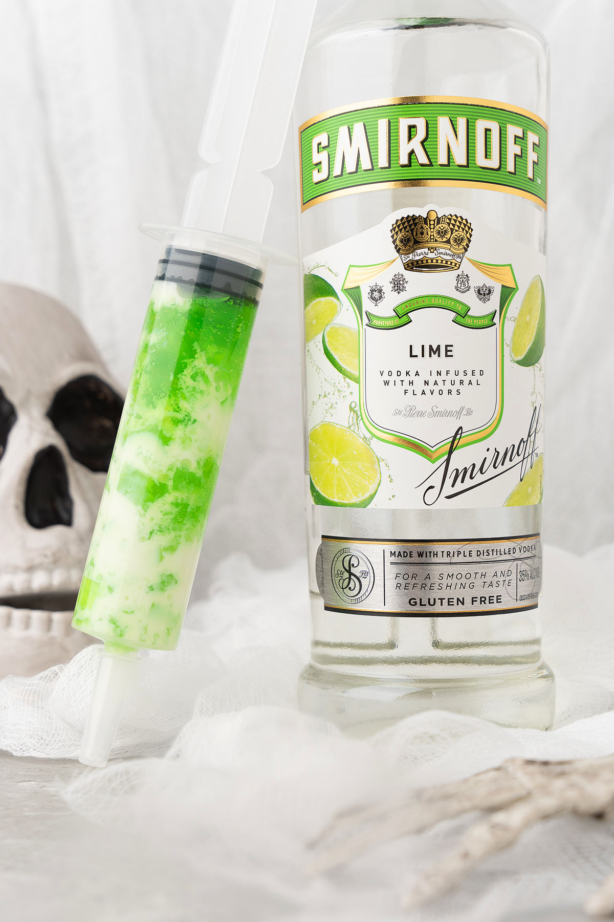 A bottle of Smirnoff Lime vodka stands next to a green-filled syringe, hinting at creative vodka syringe shots; a skull lurks in the background.