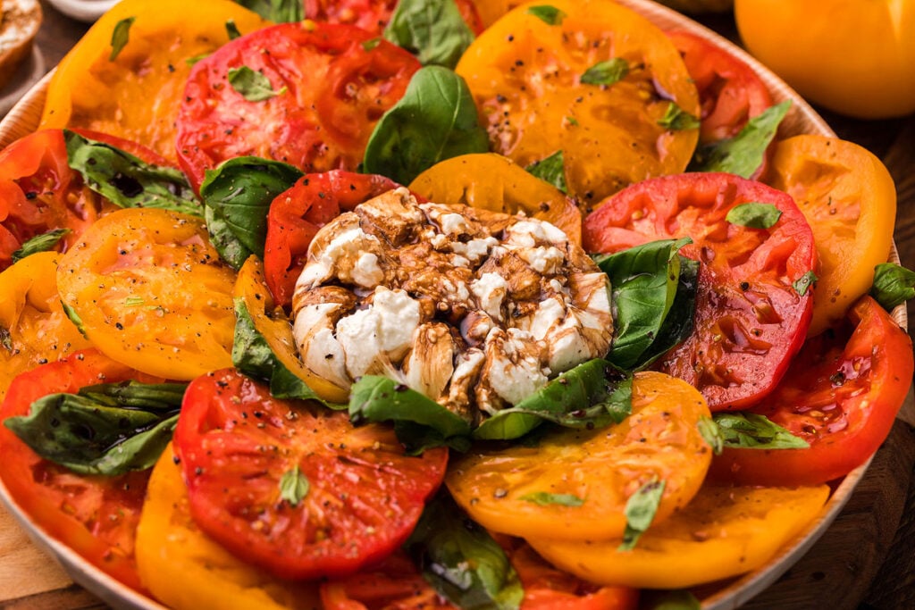 A plate of sliced red, yellow, and orange tomatoes garnished with basil leaves and topped with a whole roasted garlic bulb drizzled with balsamic glaze.