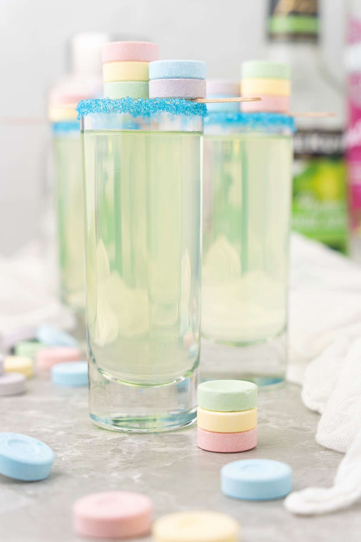 A tall glass with a blue sugar rim, pastel candies on top, and more SweeTarts candy shooters scattered around—this playful cocktail recipe features black cherry-flavored rum and sour apple schnapps for a fun, colorful treat.