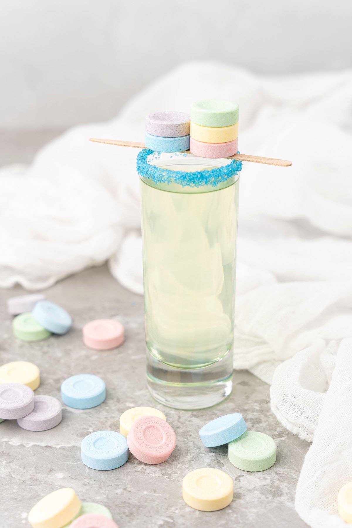 A tall glass of yellow cocktail with blue sugar rim, topped with colorful SweeTarts candy shooters and a wooden stick. Sweet and sour mix inside, candies scattered around for a fun twist.