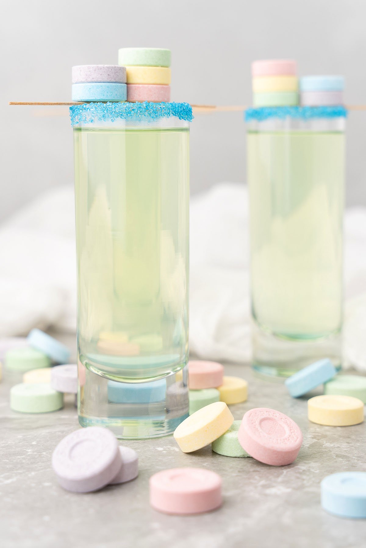 Two tall glasses with blue sugar rims, filled with a light sweet and sour mix, and topped with pastel SweeTarts candy shooters on a toothpick.