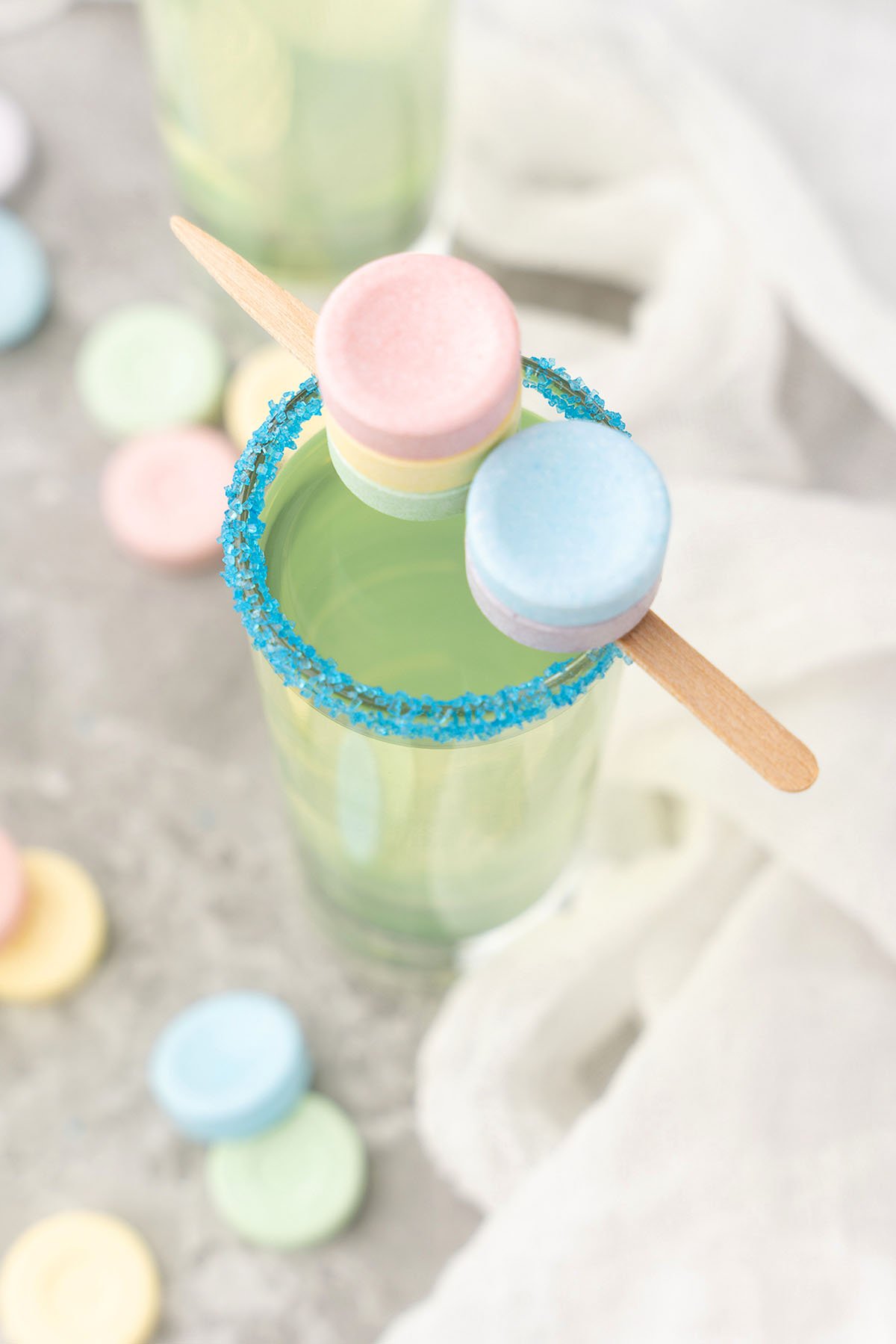 A green Sweet Tart Shot with a blue sugar rim, made with black cherry-flavored rum and sour apple schnapps, topped with pastel candy wafers on a wooden stick.