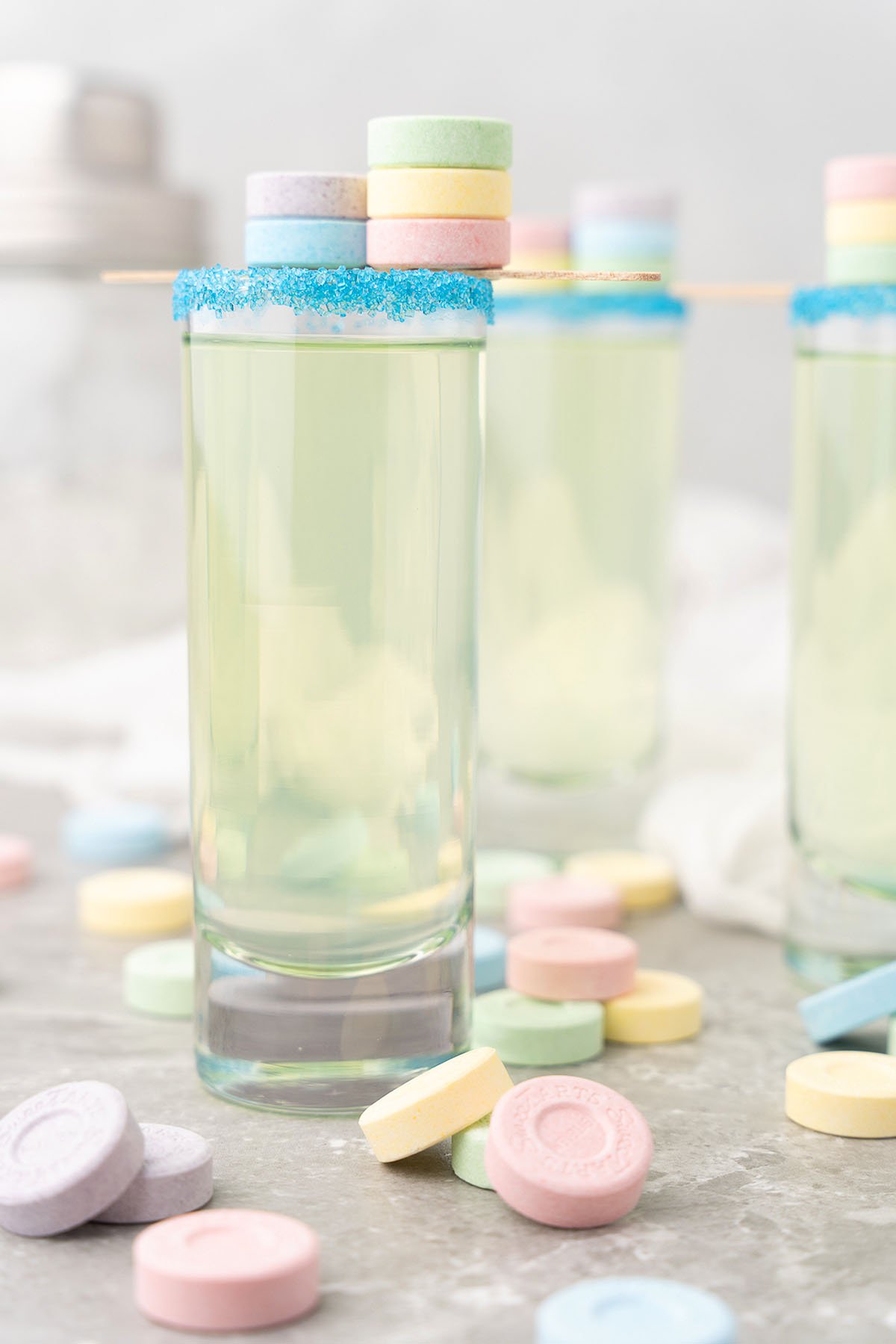 A pastel cocktail in a tall glass with a blue sugar rim and sweet and sour mix, garnished with stacked candy, surrounded by colorful SweeTarts candy shooters.