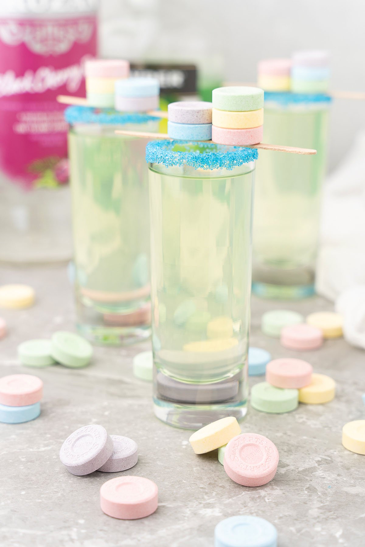 Tall glasses of Sweet Tart Shots with blue sugar rims, filled with yellow-green sweet and sour mix, each topped with pastel SweeTarts candy shooters and surrounded by colorful candies.