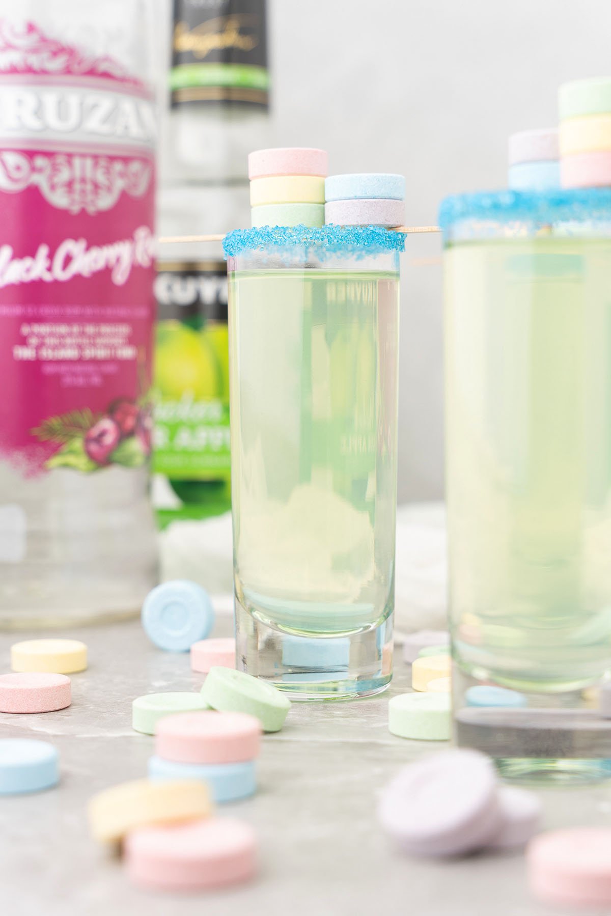 Two pastel-colored shots with blue sugar rims, featuring black cherry-flavored rum and sour apple schnapps, garnished with candy and surrounded by sweet tarts and bottles—an irresistible cocktail recipe for any occasion.