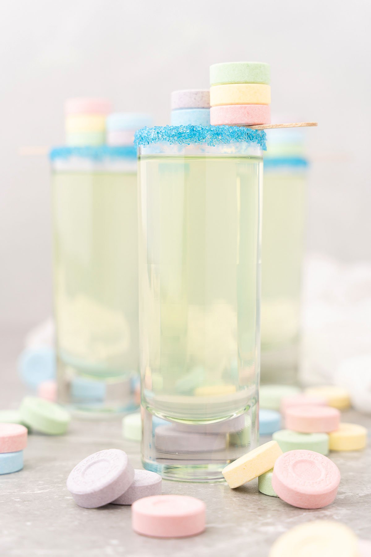 A tall glass with a blue sugar rim, filled with a clear drink and topped with pastel SweeTarts candy shooters on a stick, perfect for a colorful sweet and sour mix cocktail recipe.