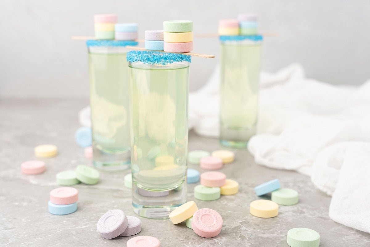 Three yellow-green SweeTarts candy shooters in shot glasses, made with black cherry-flavored rum and sour apple schnapps, are garnished with blue sugar and candy, surrounded by pastel sweets—a playful cocktail recipe for any party.