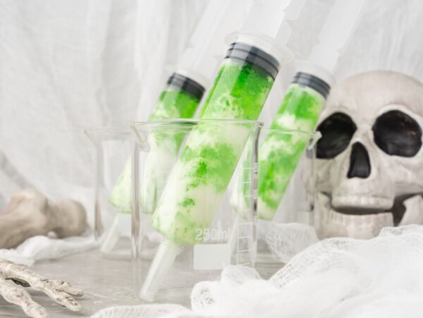 Green liquid-filled syringes in beakers hint at Ectoplasm Syringe Vodka Jello Shots, with a skull and skeleton hand on a white, spooky background—perfect for Halloween jello shots with a twist of Smirnoff lime vodka.