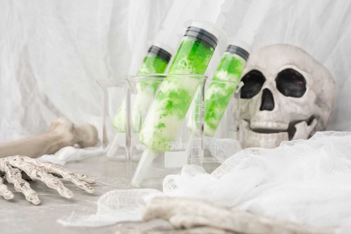 Green syringes filled with Ectoplasm Syringe Vodka Jello Shots rest in beakers beside a fake skull and skeleton hand, with white gauze in the background—perfect for your spooky party when you want to make jello shots in syringes.