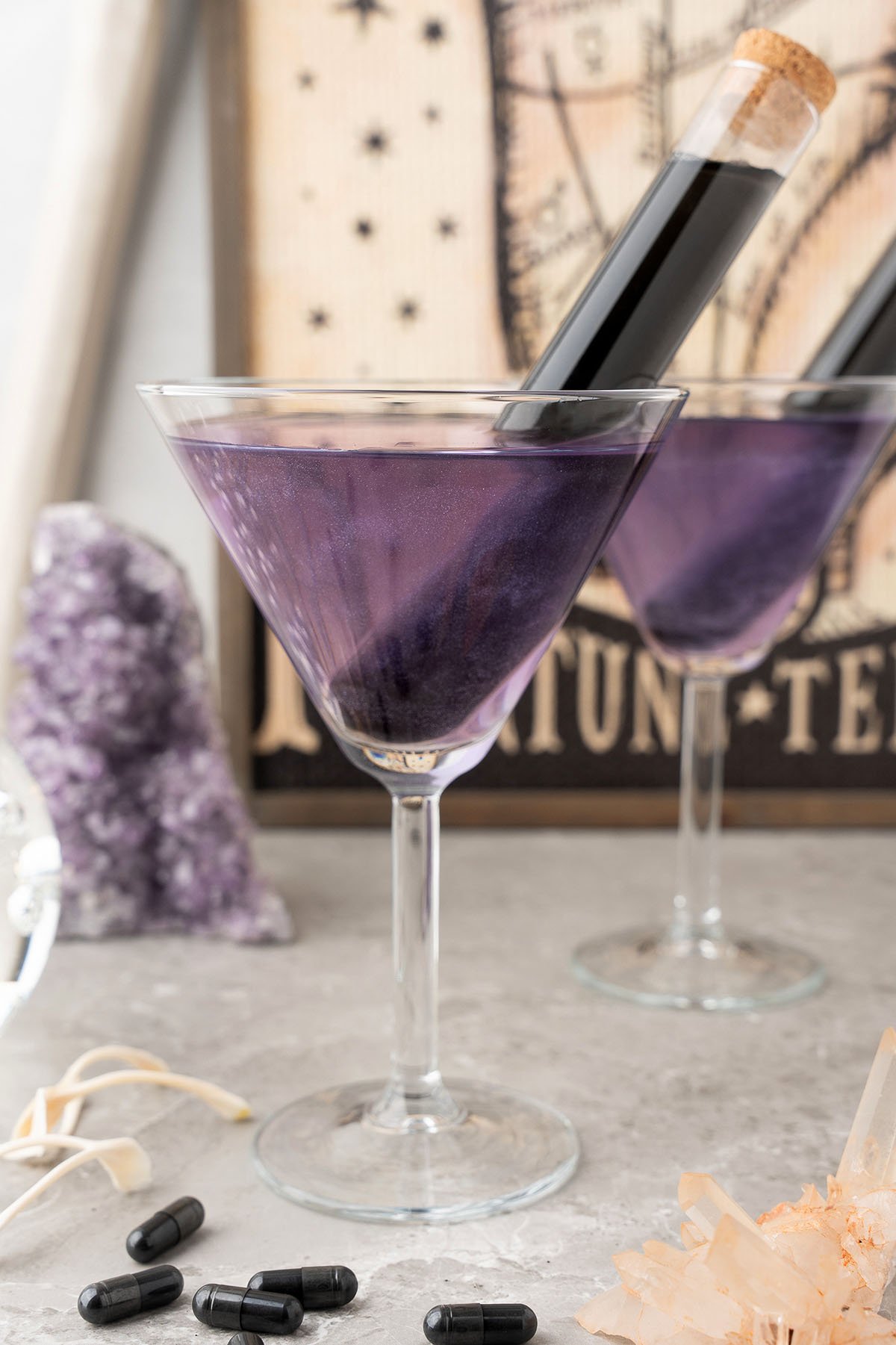 A spooky alcoholic purple cocktail in a martini glass with a vial inside, surrounded by crystals and black capsules, featuring sour grape schnapps and strawberry rum for a perfectly themed mixed drink.