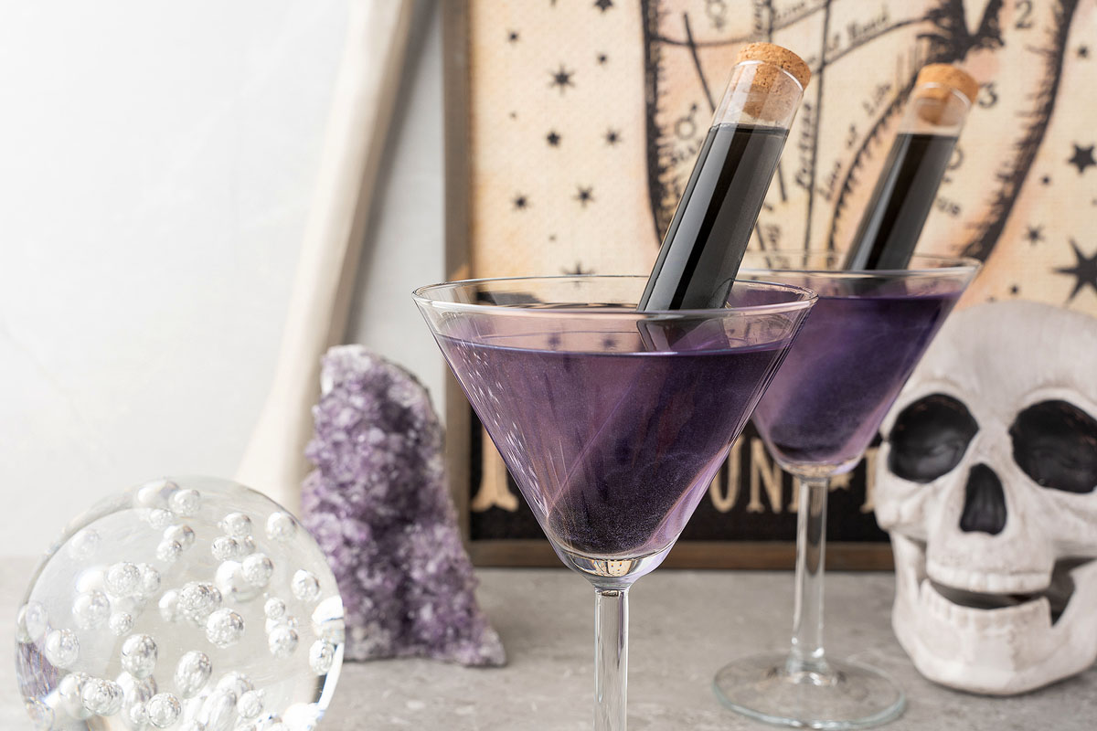 Two martini glasses filled with a purple drink, each containing a corked test tube, are set on a marble surface. The background features a decorative piece with a celestial design, a piece of amethyst, a crystal ball, and a skull prop.