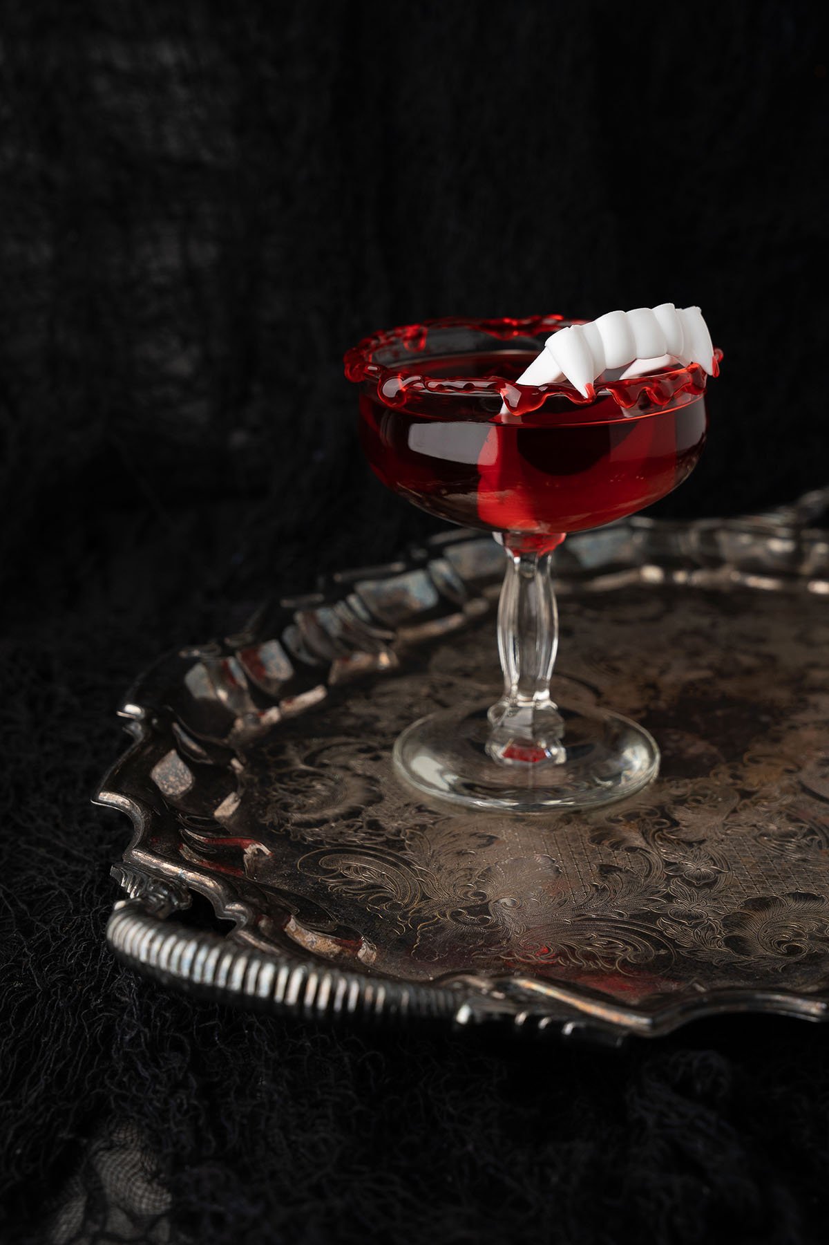A red Cursed Immortal Halloween Rum Cocktail with fake vampire teeth sits in a glass on a vintage silver tray, ready to haunt your party as the ultimate black cherry rum beverage against a dark backdrop.