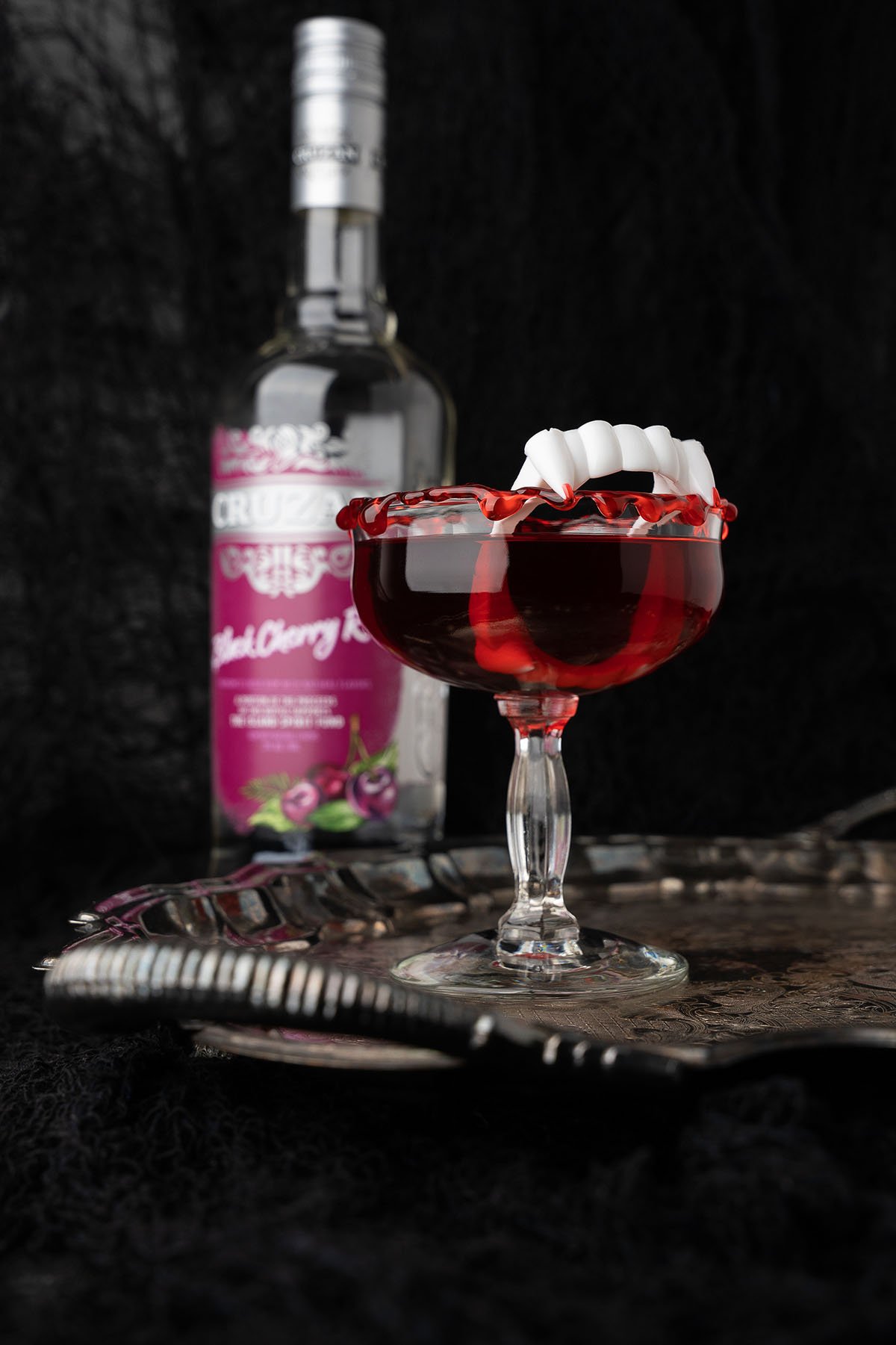 A blood red cocktail with gummy teeth garnish in a glass, featuring a bottle of cherry vodka in the background—perfect for serving as your Cursed Immortal Halloween Rum Cocktail.