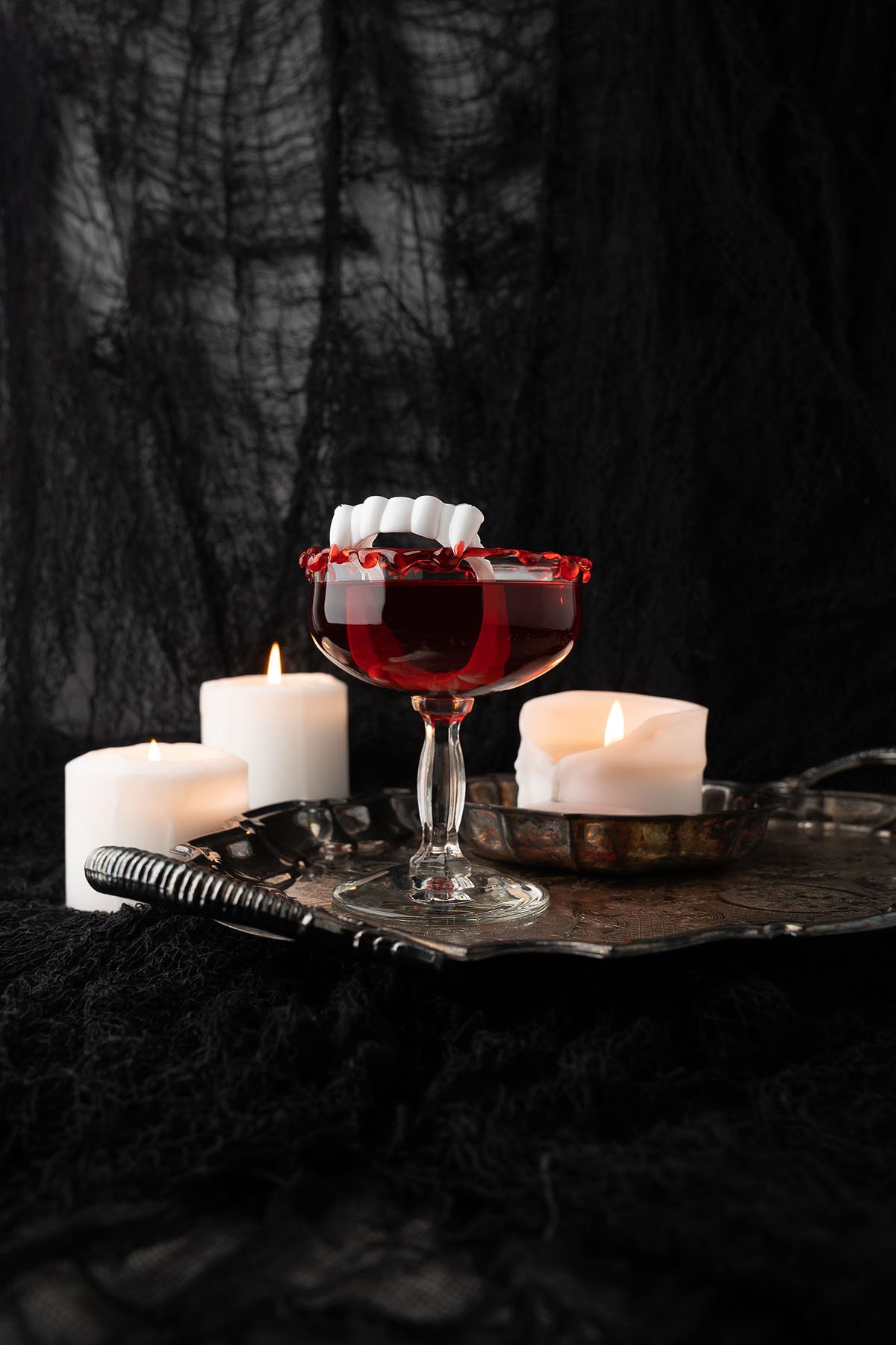 A blood red cocktail made with black cherry rum sits on a tray, adorned with fake vampire teeth and surrounded by lit white candles and black fabric.