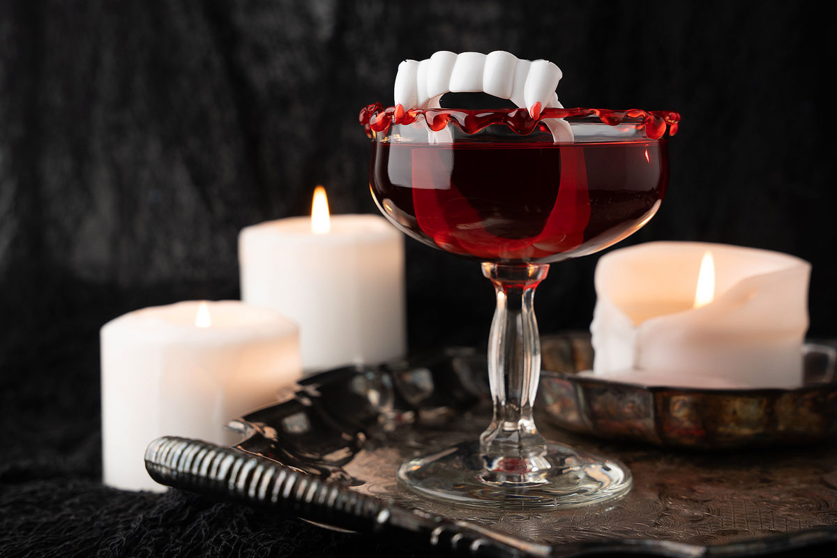 A spooky-themed cocktail in a glass is topped with fake vampire fangs and rimmed with red syrup. The glass is set against a dark, textured background alongside three lit white candles, creating a Halloween ambiance. A vintage tray and assorted utensils are visible.