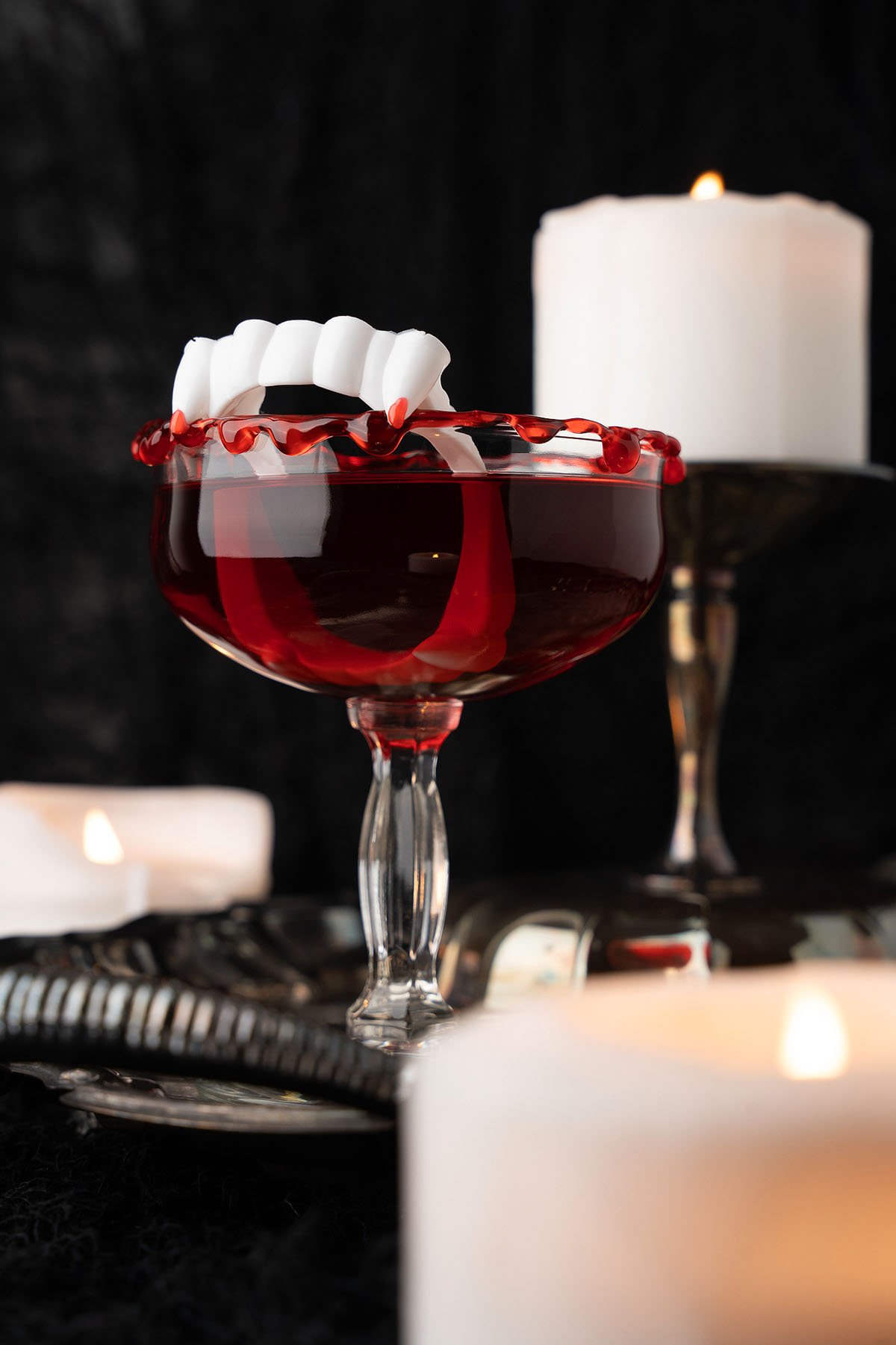 A red Cursed Immortal Halloween Rum Cocktail in a glass with fake vampire teeth, surrounded by lit white candles.