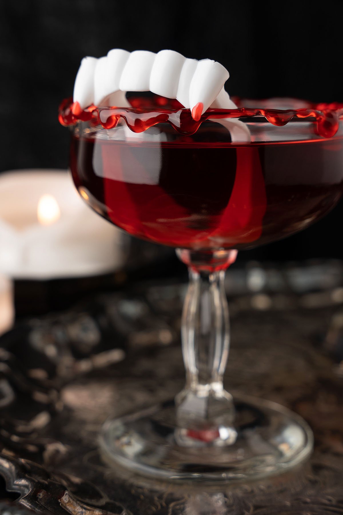 A red cocktail in a glass with fake vampire fangs on the rim, dripping “blood” syrup—this black cherry rum Halloween drink makes a spooktacular party beverage.