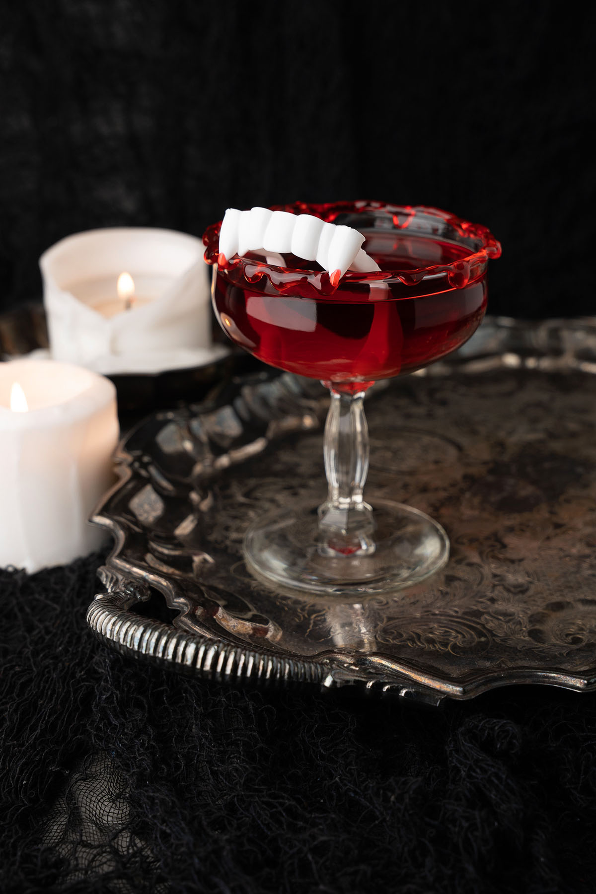 A blood red cocktail with a fake teeth garnish sits on a tray beside lit white candles, set against a black background. This Cursed Immortal Halloween Rum Cocktail features bold flavors and eerie vibes, perfect for spooky celebrations.