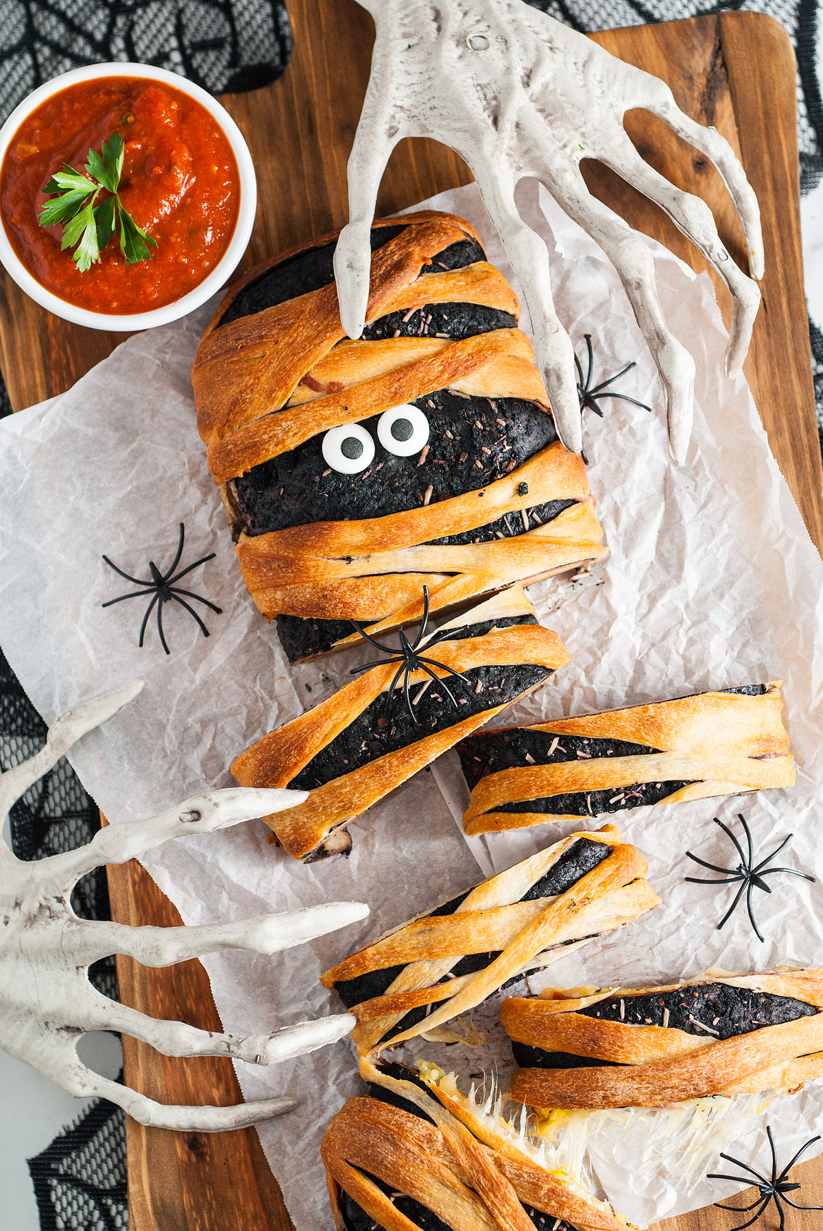 A Halloween Mummy Stromboli Dinner made with Pillsbury refrigerated pizza dough, wrapped like a mummy with candy eyes and surrounded by fake skeleton hands and spiders for the perfect Halloween themed food display.