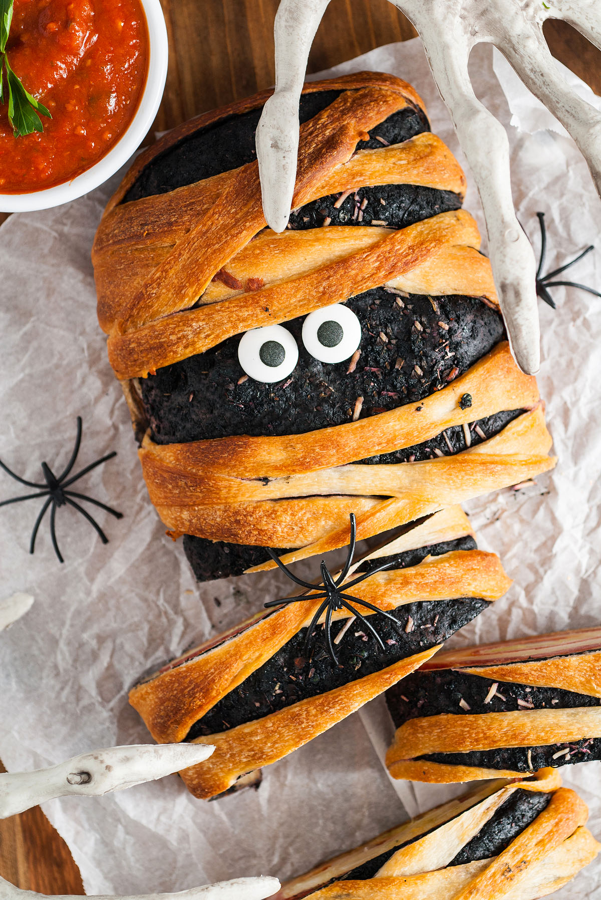 A mummy-shaped bread loaf made with Pillsbury refrigerated pizza dough and candy eyes, wrapped in golden pastry—perfect as a spooky Halloween dinner or appetizer, and surrounded by fake spiders and bones.