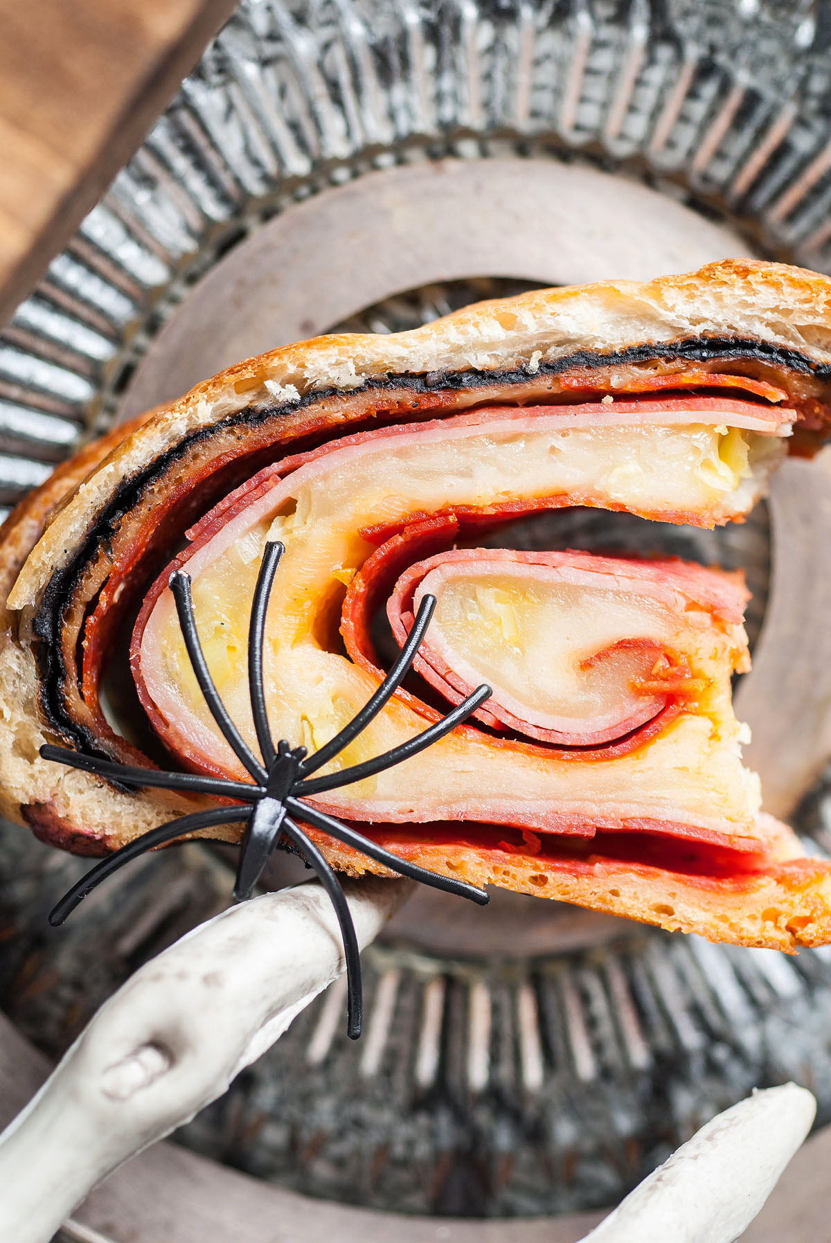 A slice of rolled sandwich with layers of meat and cheese, topped with a fake black spider for spooky fun—a perfect Halloween themed food for your dinner or appetizer table.
