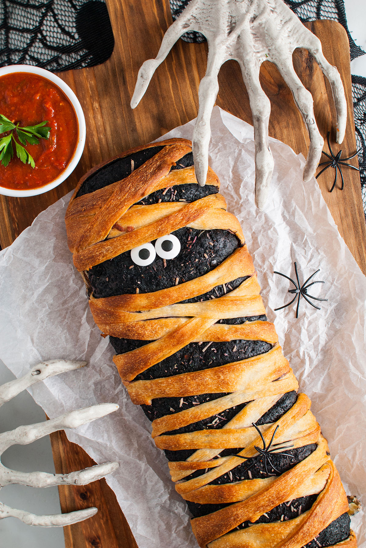 A loaf of bread made from Pillsbury refrigerated pizza dough is decorated like a mummy with googly eyes, fake spider props, and skeleton hands nearby for a fun Halloween themed food.