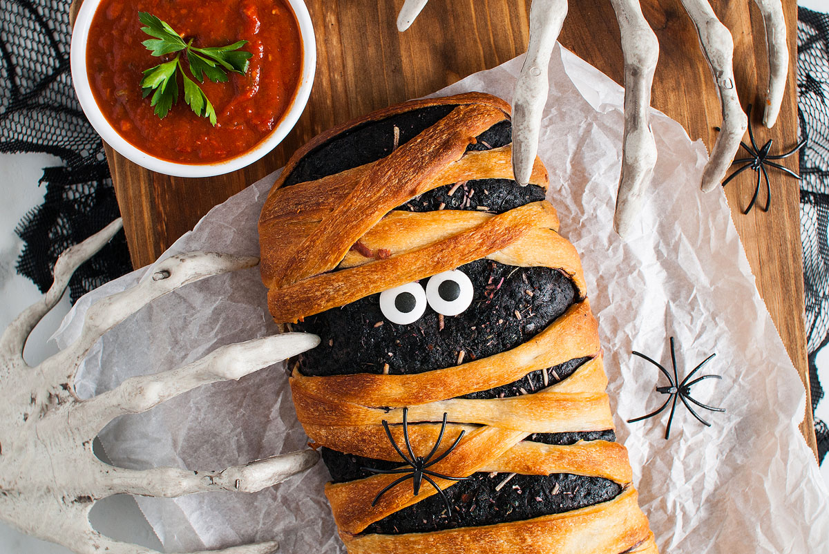 A loaf of bread made from Pillsbury refrigerated pizza dough is decorated as a mummy with candy eyes, plastic spiders, and a bowl of red sauce nearby—perfect for a spooky Halloween dinner or appetizer.