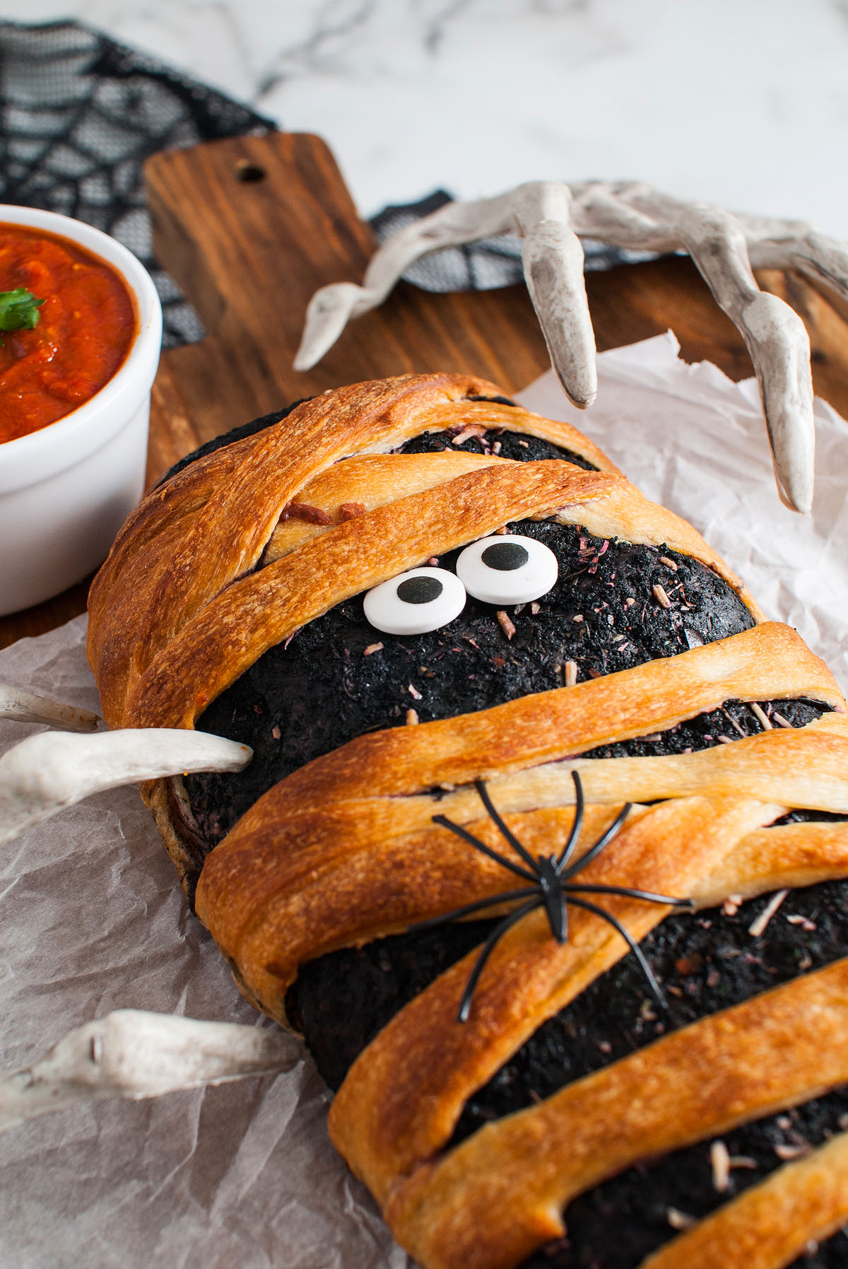 A Halloween themed food: homemade stromboli wrapped like a mummy with candy eyes, fake spider, and skeleton hands, made using Pillsbury refrigerated pizza dough, served next to tomato sauce.