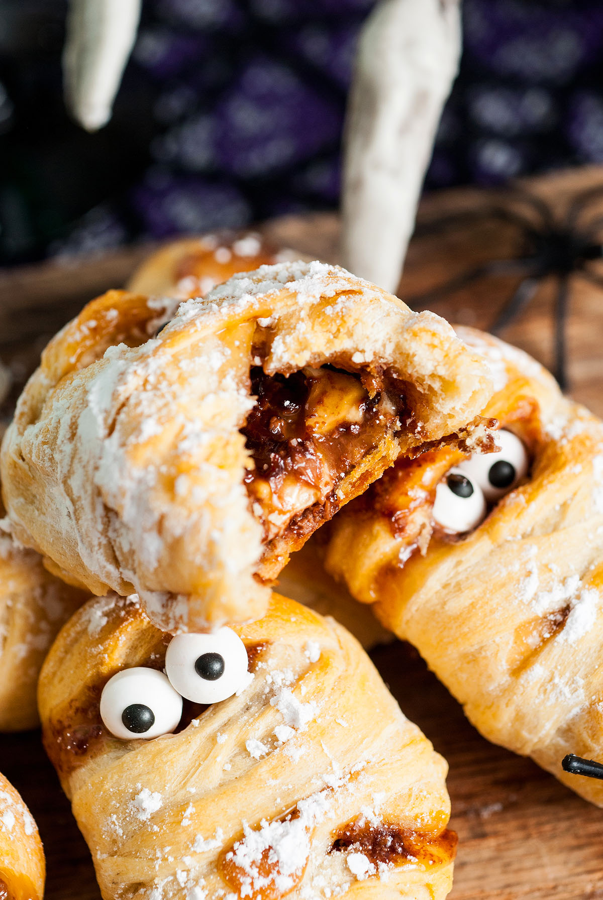 Three spooky pastries decorated like mummies with candy eyes and powdered sugar on top, one broken open to reveal a delicious fun size Snickers filling.
