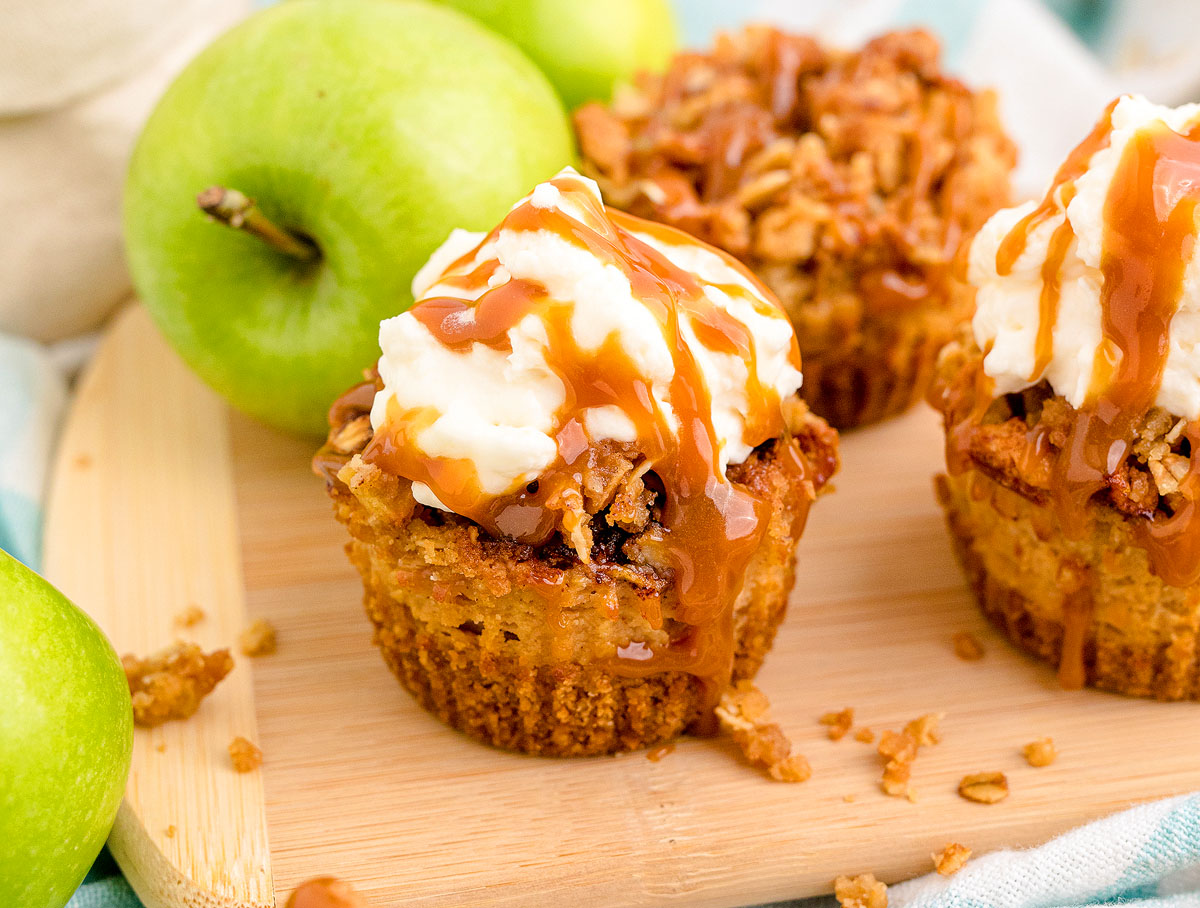 Close-up of two apple crumble cupcakes topped with whipped cream and caramel sauce on a wooden board. The cupcakes have a golden-brown crumbly texture, and green apples are in the background, suggesting the flavor and main ingredient.