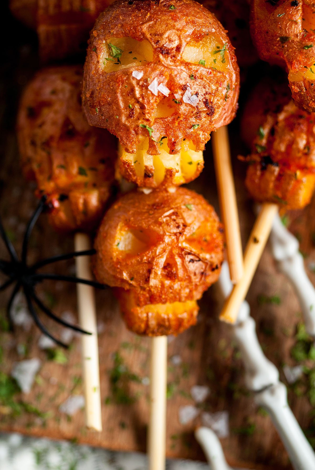 Skull-shaped fried snacks on sticks, resembling Spooky Halloween Skull Potatoes, are garnished with herbs and salt, with a black plastic spider nearby.