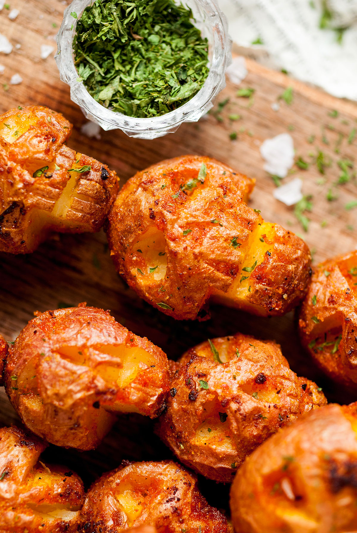 Close-up of crispy roasted potatoes on a wooden board, styled as Spooky Halloween Skull Potatoes, with a small bowl of chopped herbs nearby—perfect as a shrunken head-inspired Halloween side dish or appetizer.