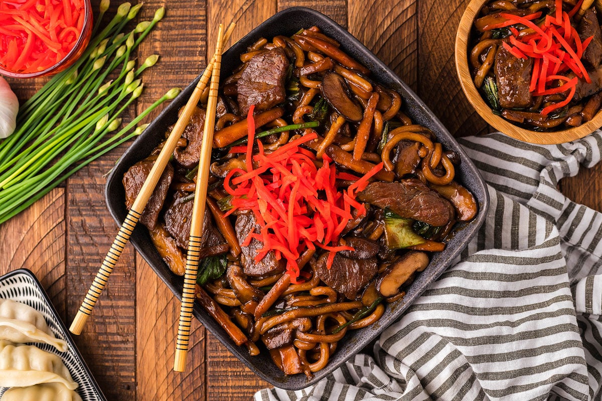 A bowl of stir-fried noodles with beef, vegetables, and sliced mushrooms, garnished with shredded red pickled ginger. A pair of chopsticks rests on the rim of the bowl. Surrounding items include a striped cloth napkin, green onions, garlic, and dumplings.