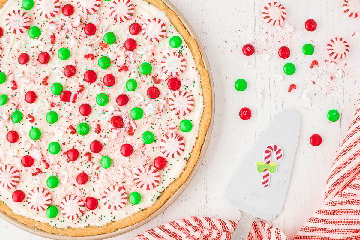 Large cookie made from refrigerated sugar cookie dough, topped with white frosting, festive red and green candy toppings, and peppermint mints—perfect as a Christmas dessert pizza on a white table.
