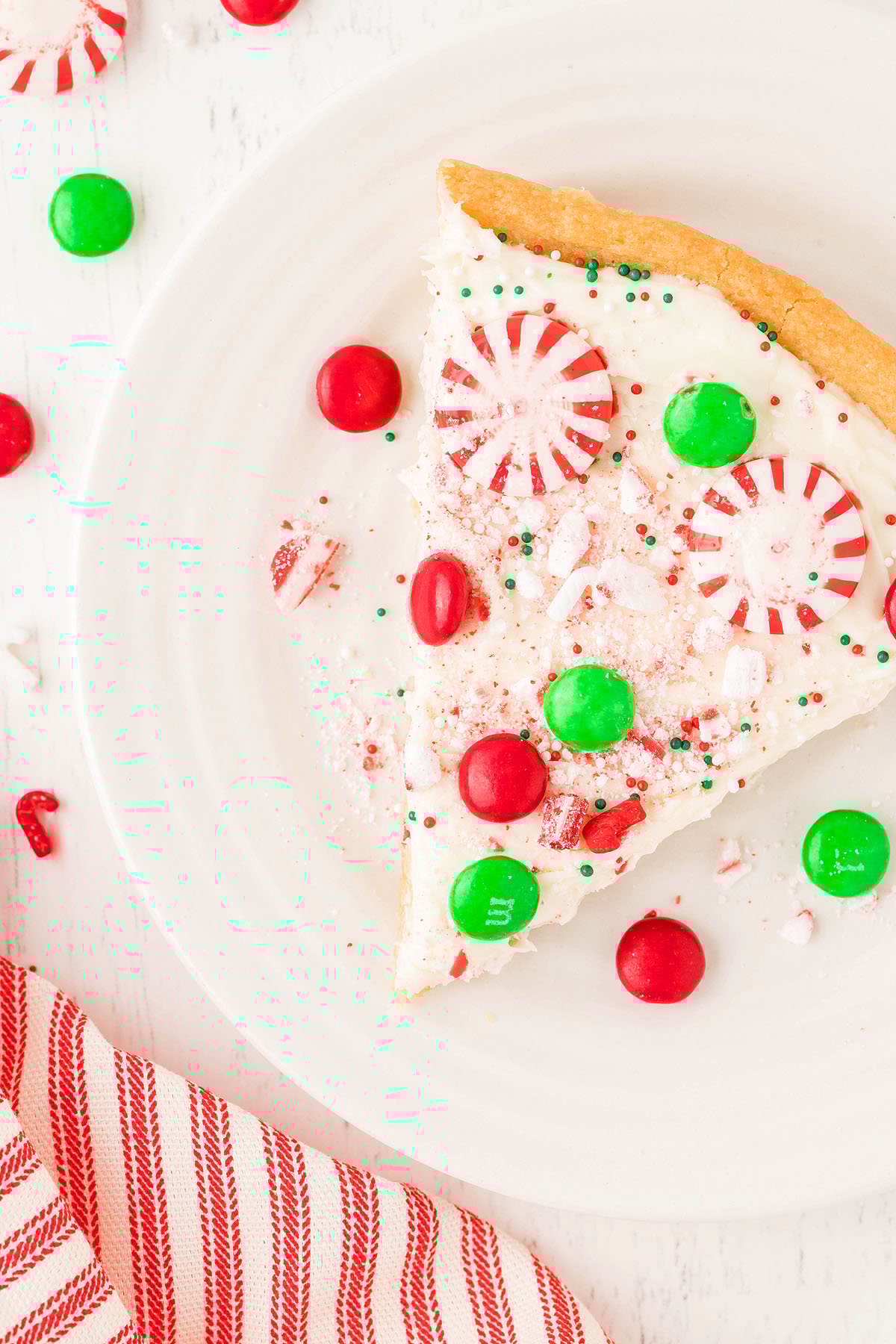 A slice of Christmas dessert pizza made with refrigerated sugar cookie dough, topped with white frosting, red and green candies, and peppermint pieces.
