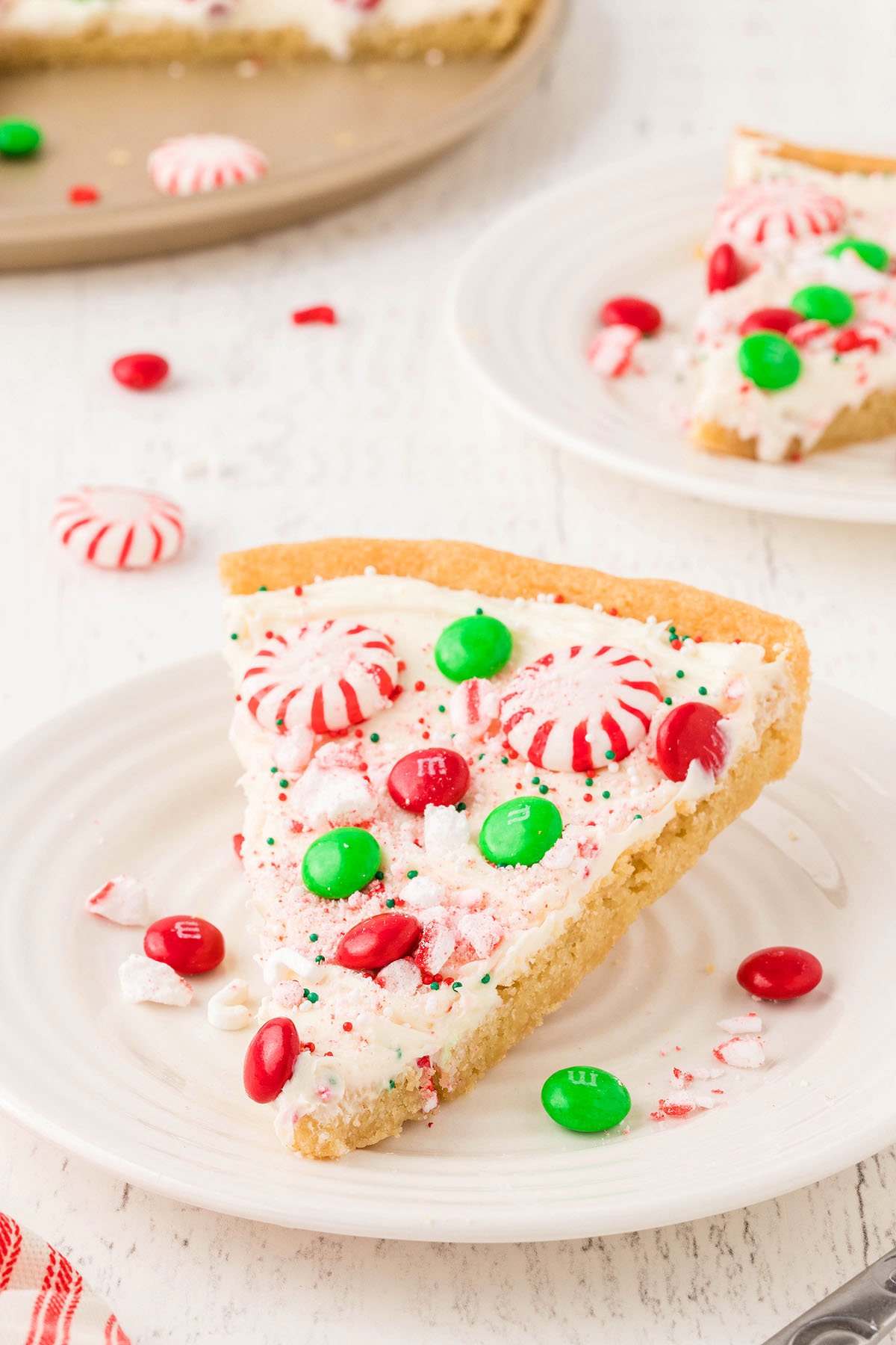 A slice of Christmas dessert pizza made with refrigerated sugar cookie dough, topped with white frosting, red and green candies, and peppermint candies on a white plate.