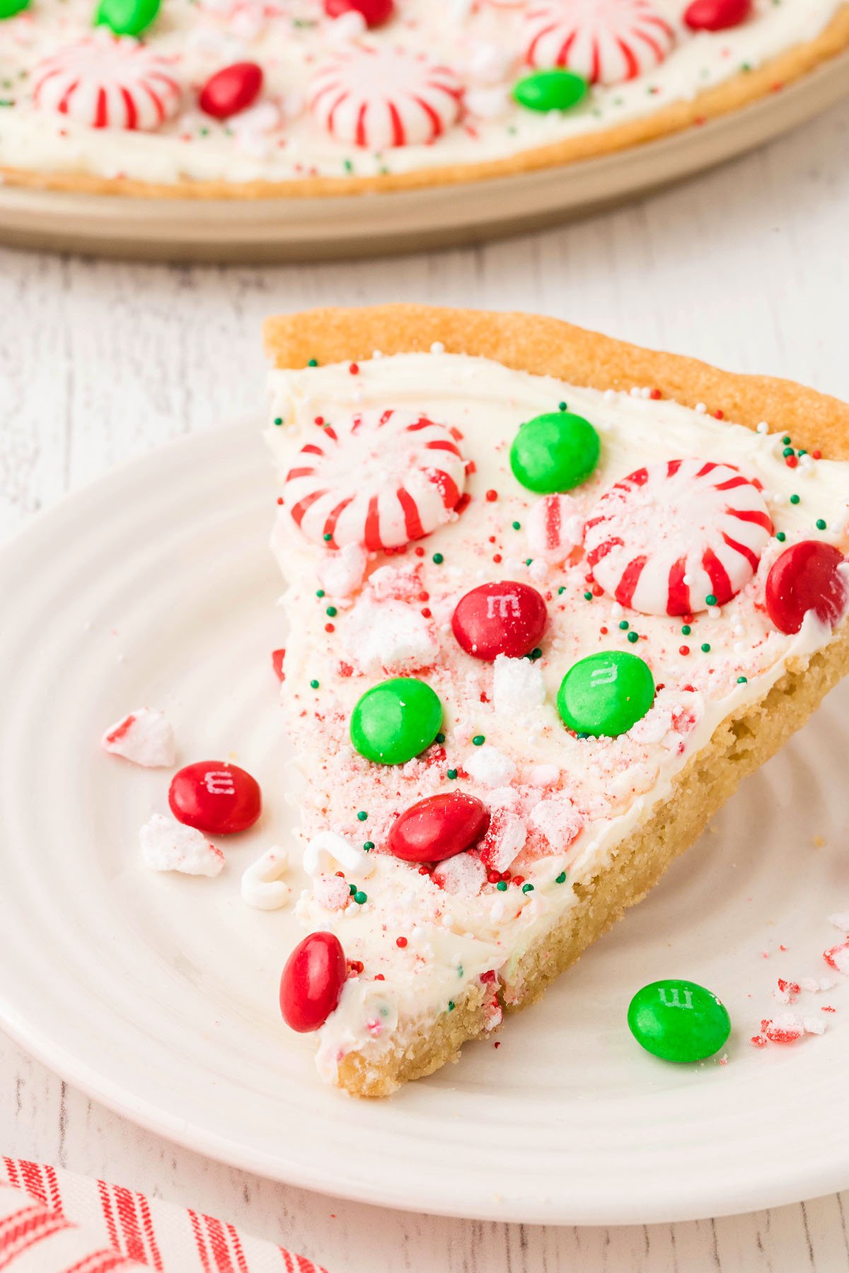 A slice of Holiday Cookie Pizza topped with white frosting, festive candy toppings, red and green candies, and peppermint candies.