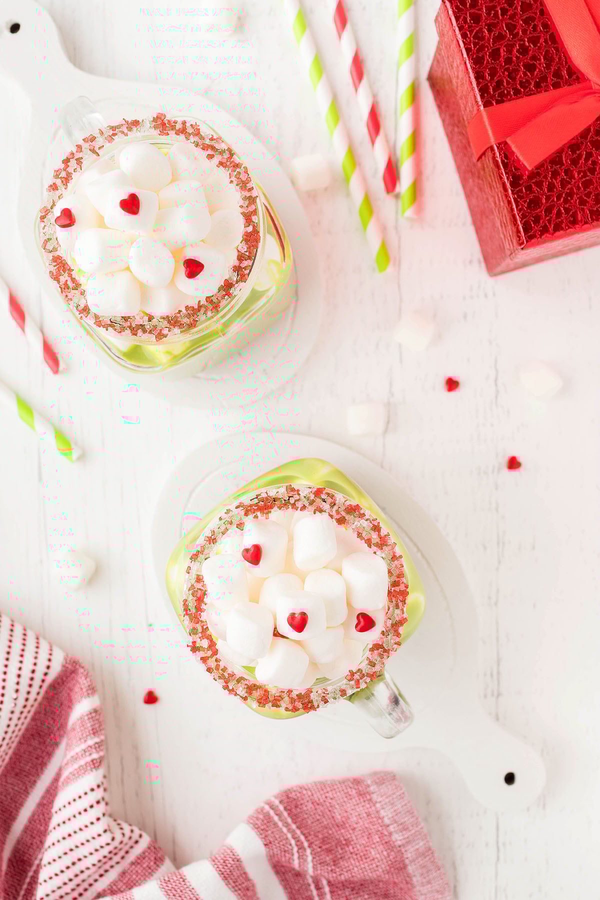 Two mugs of Grinch Hot Chocolate with marshmallows and red candy hearts sit beside festive straws and a red gift box.