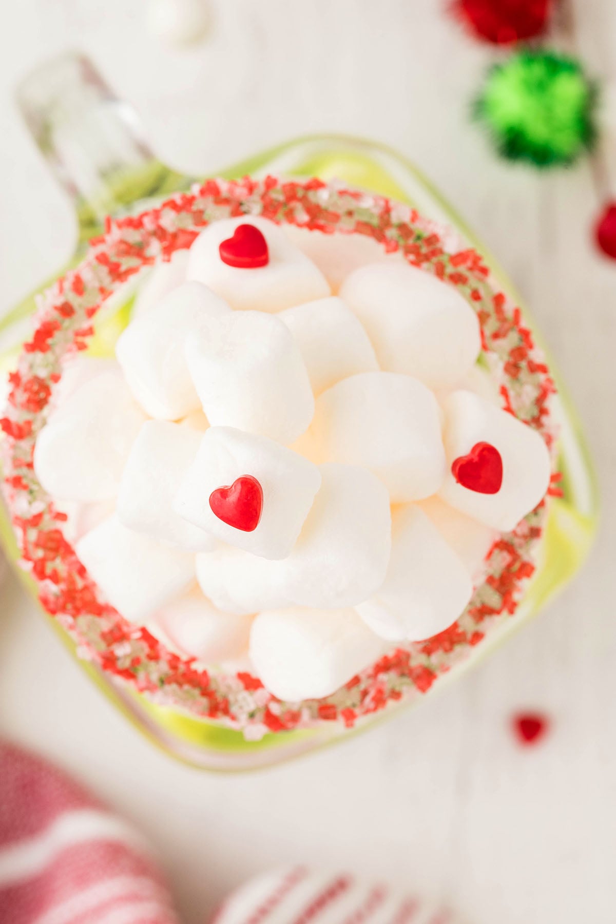 A fun holiday party drink, this Green Grinch hot chocolate is served in a mug of marshmallows topped with red candy hearts and rimmed with festive red and green sprinkles.